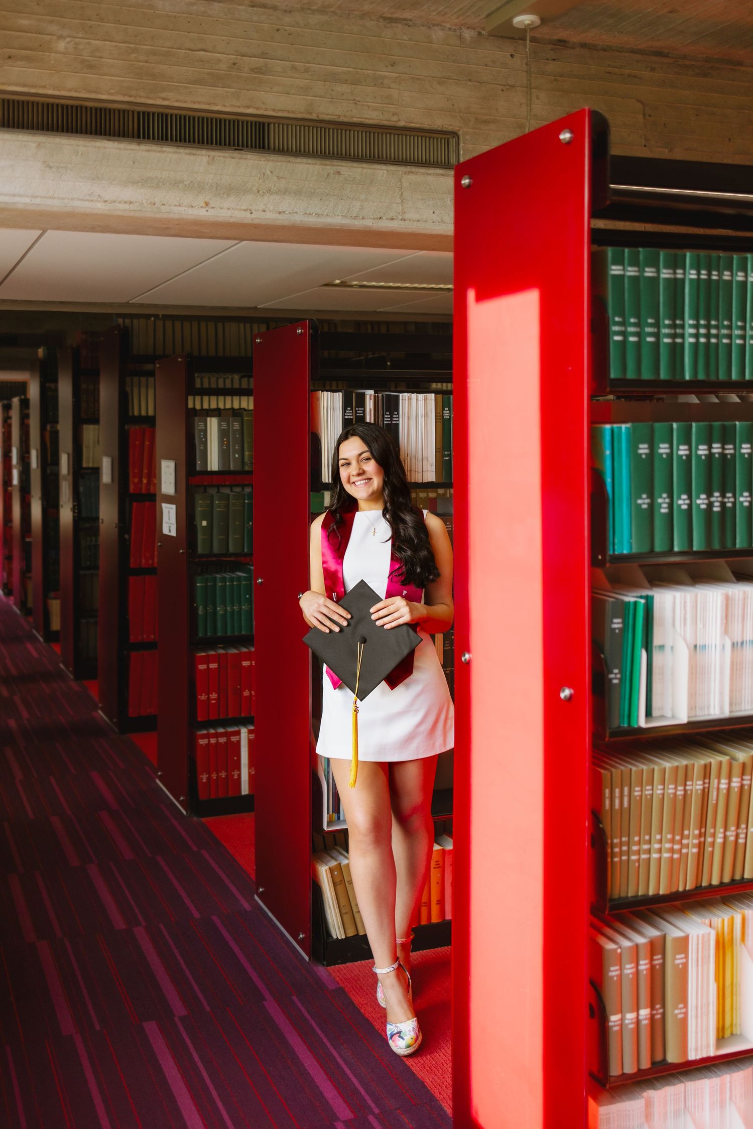 College grad holds a book while standing between bookshelves in a library with red shelving units and bound volumes at UMASS Dartmouth MA