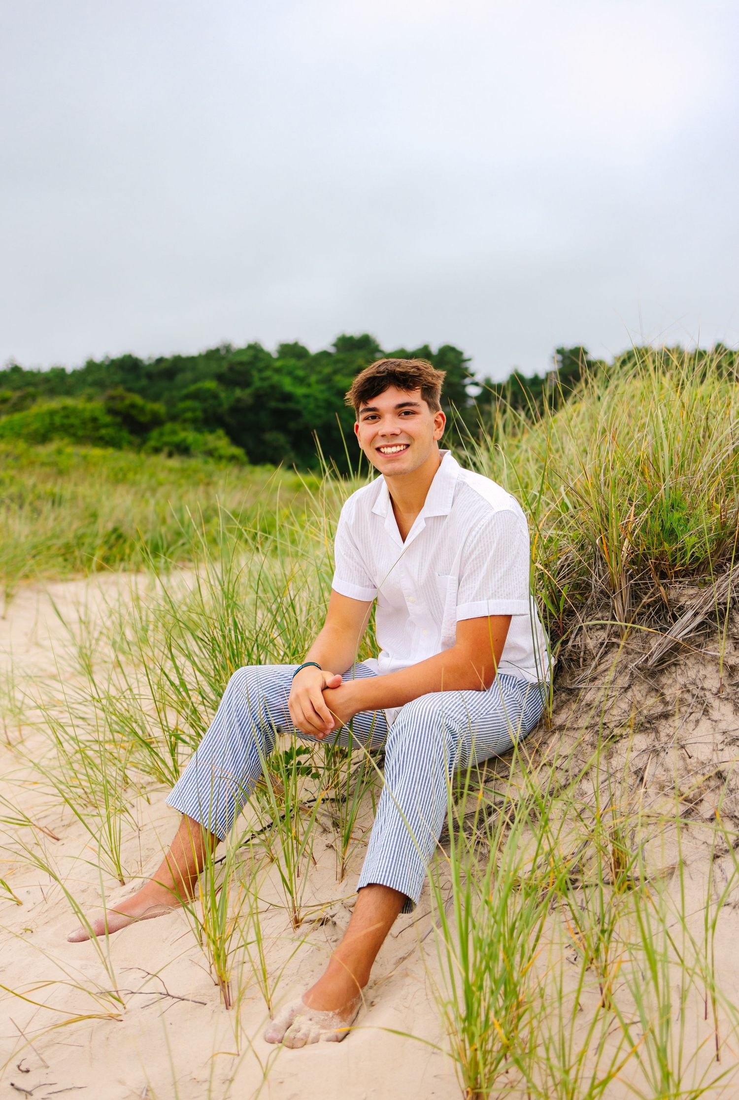 A high school senior sits on sandy dunes with tall grass on a cloudy summer day at Horseneck Beach in Westport Massachusetts