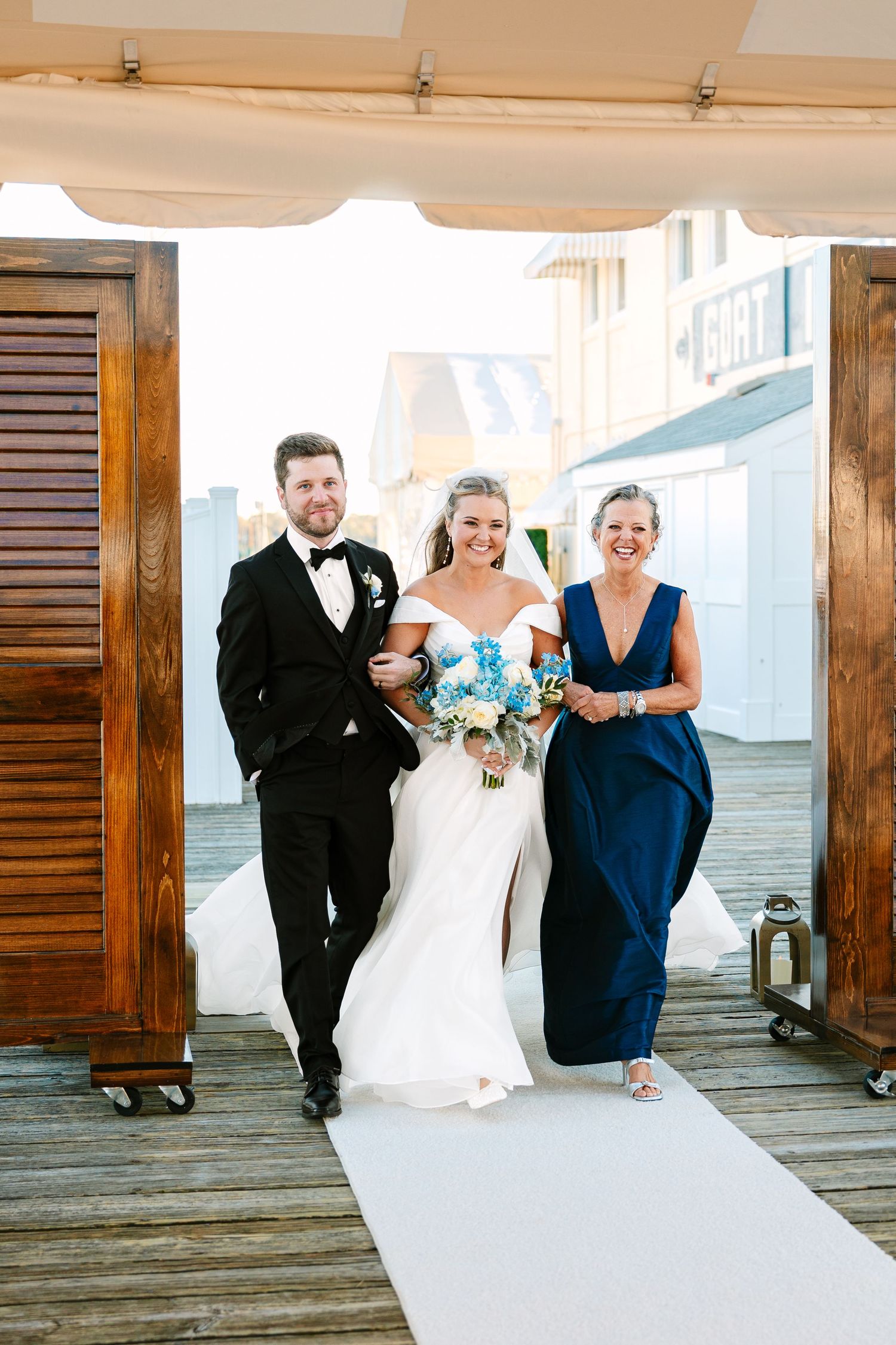 Bride entering wedding ceremony walking with family on a wooden boardwalk with white carpet runner and wooden shutters in background in RI