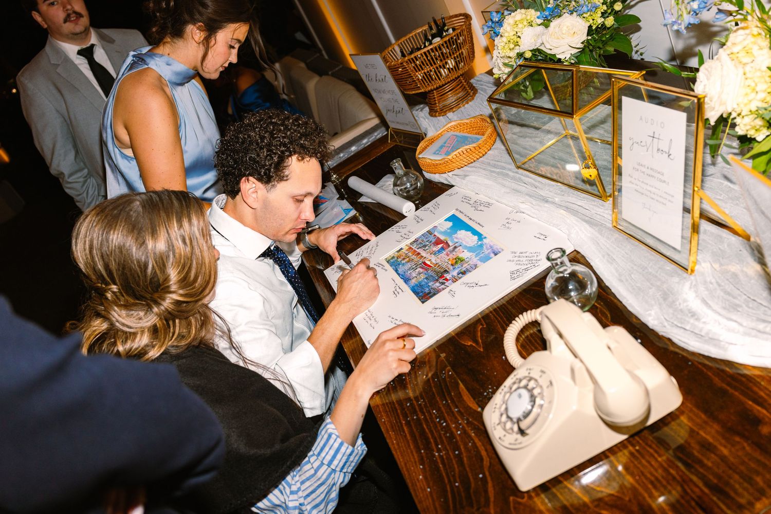 Guests gather around a wooden table at an event looking at photo albums while a vintage white telephone sits nearby.