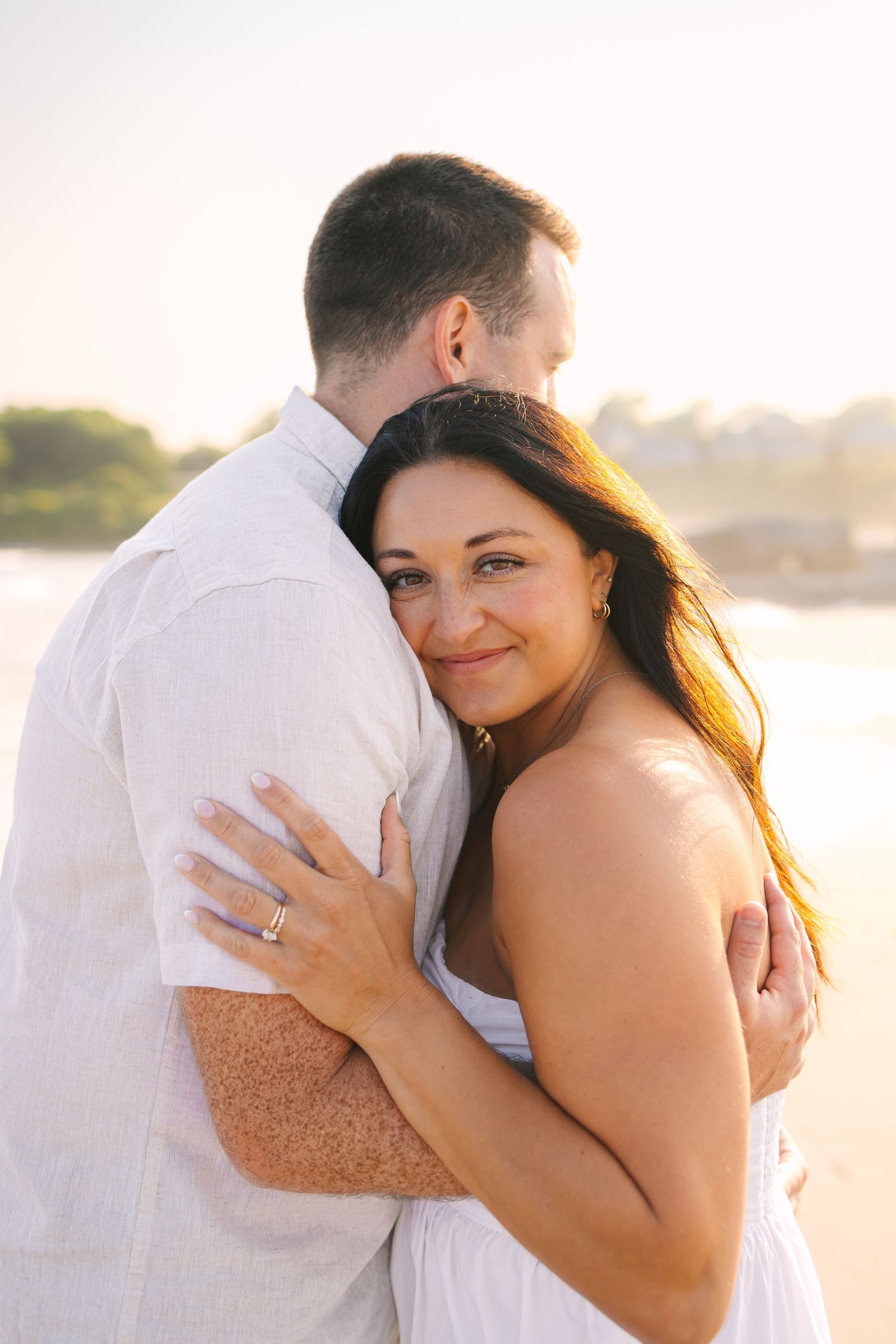 A couple embraces on the beach at sunset wearing white clothing while showing off an engagement ring at Second Beach in Newport Rhode Island