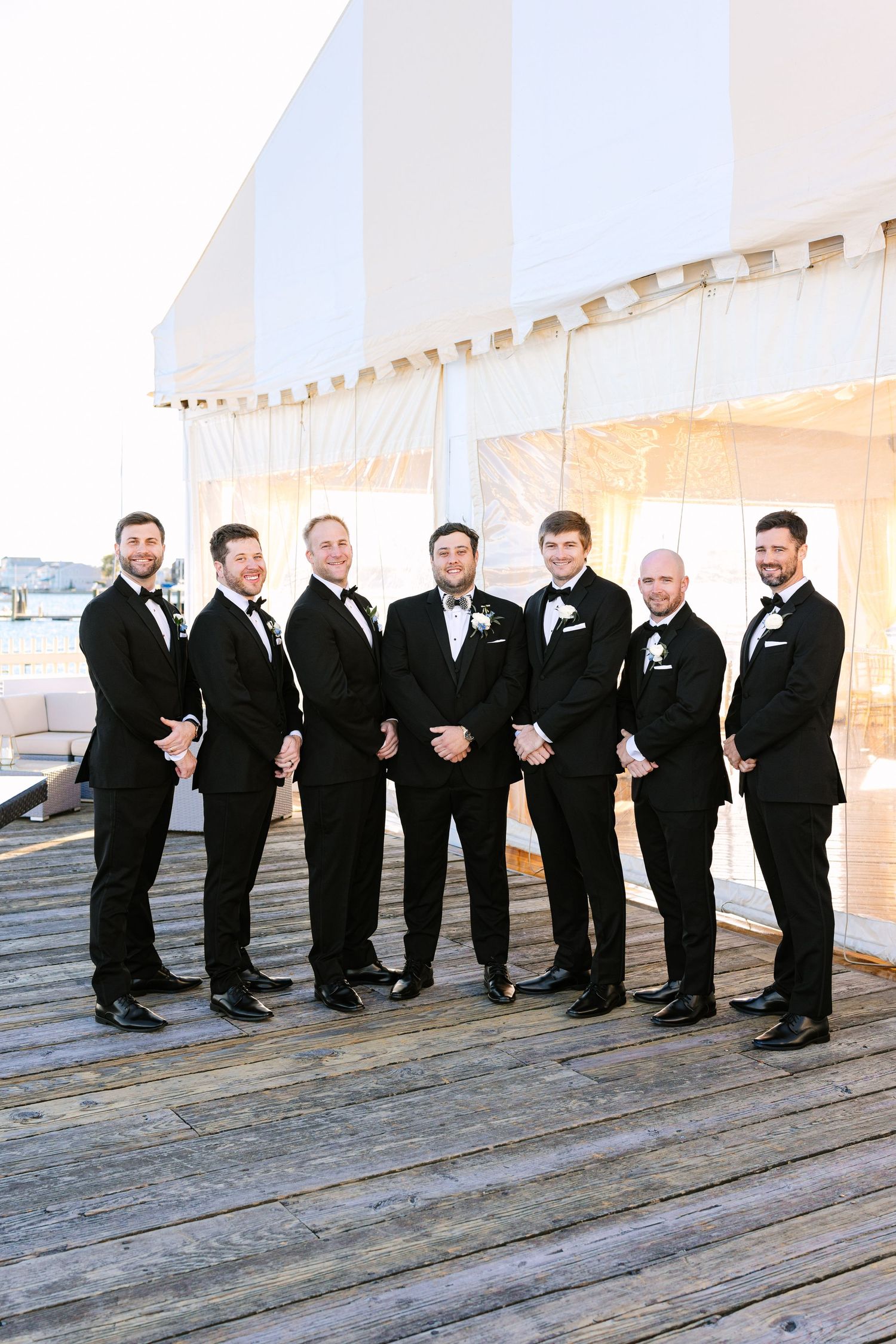 A group of groomsmen in black tuxedos pose playfully on wooden deck boards at a wedding venue.