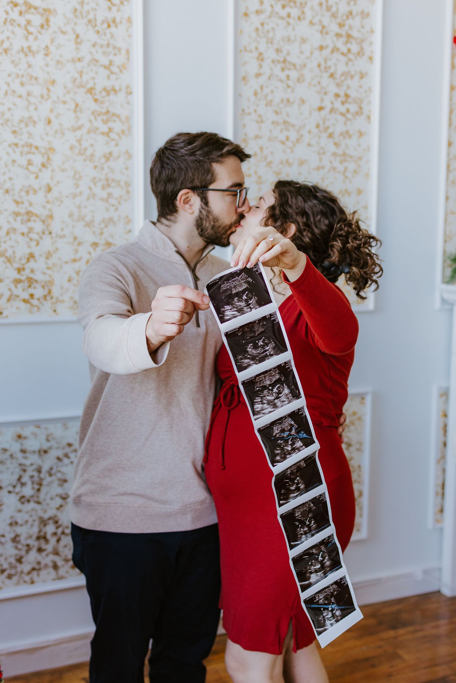 Couple in casual attire sharing a kiss while holding ultrasound photos in a Christmas themed room for maternity photos in South Coast MA
