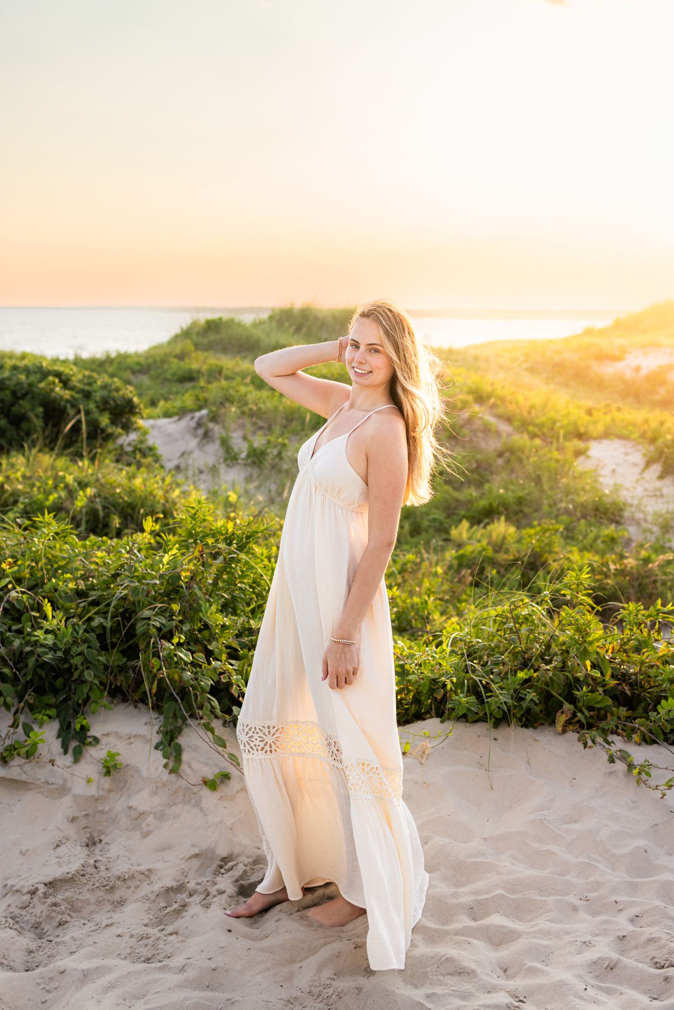 A high school senior stands by the sand dunes at sunset golden hour for senior photos at Horseneck Beach in Westport, Massachusetts