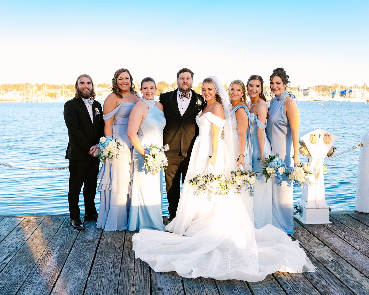 A wedding party in light blue bridesmaid dresses and formal attire poses on a wooden dock by the waterfront.