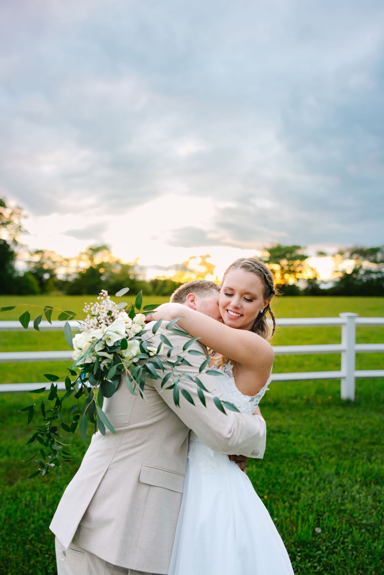 A bride and groom embrace during sunset on a countryside lawn with white fencing and gorgeous greenery at backyard wedding in Connecticut