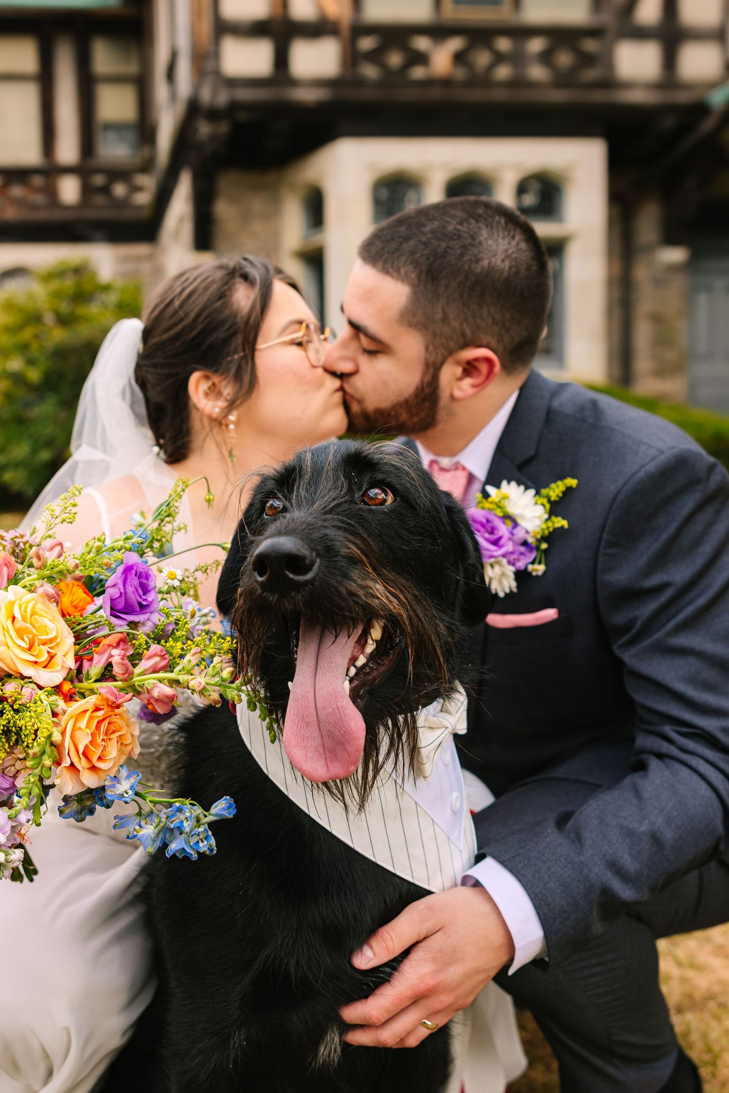 Bride and groom kissing behind their black dog wearing a suit collar, with colorful bridal bouquet outside wedding venue in South Coast MA