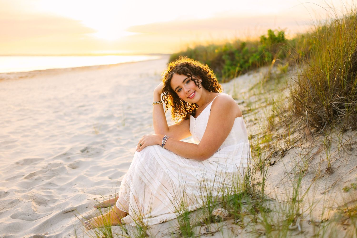A high school senior sitting by the sand dunes during sunset golden hour for senior portraits at Horseneck Beach in Westport, MA