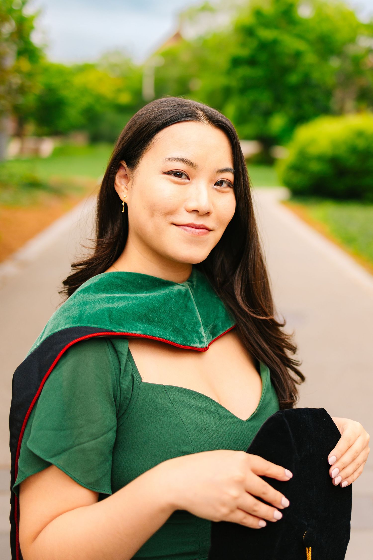 A college grad smiling outdoors on a paved pathway with lush greenery in the background for grad photos at Brown University in Rhode Island