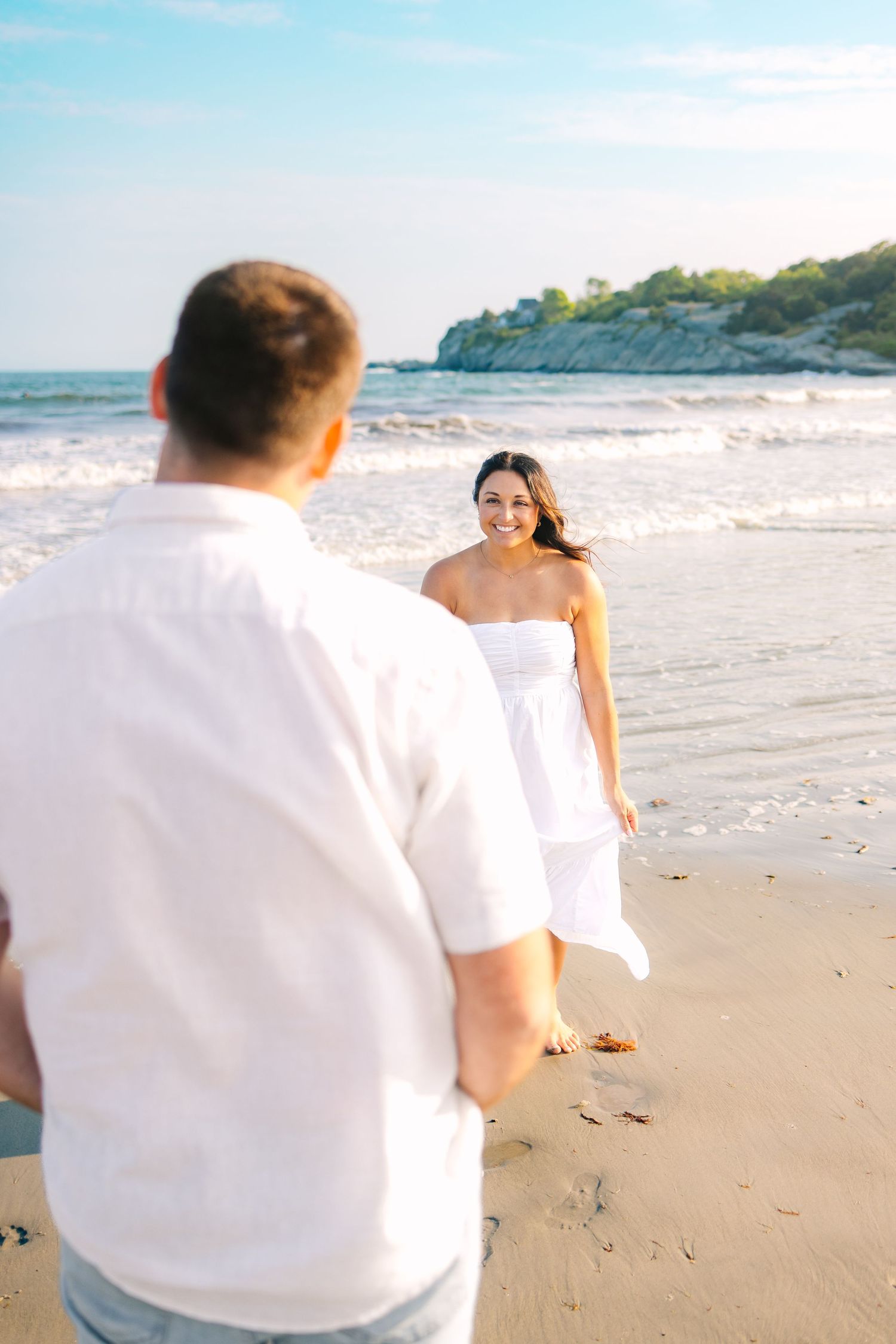 A couple in white outfits shares a romantic moment on a sunny beach with cliffs in the background at Second Beach in Newport Rhode Island