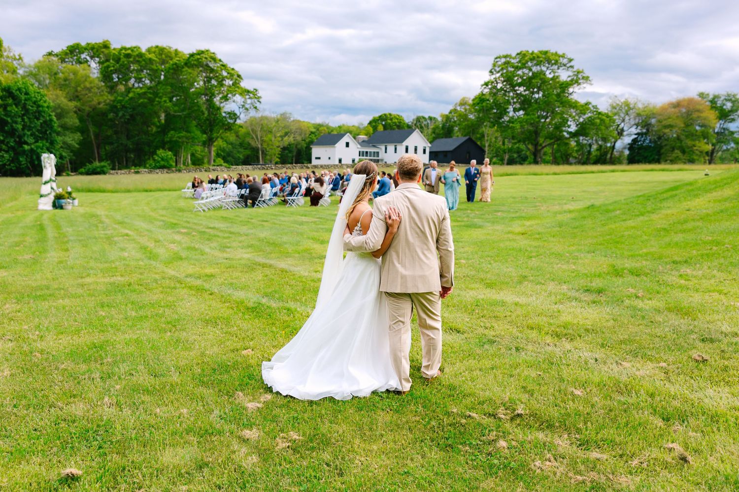 A couple in wedding attire stands in a green field with guests gathered in the background on a cloudy day.