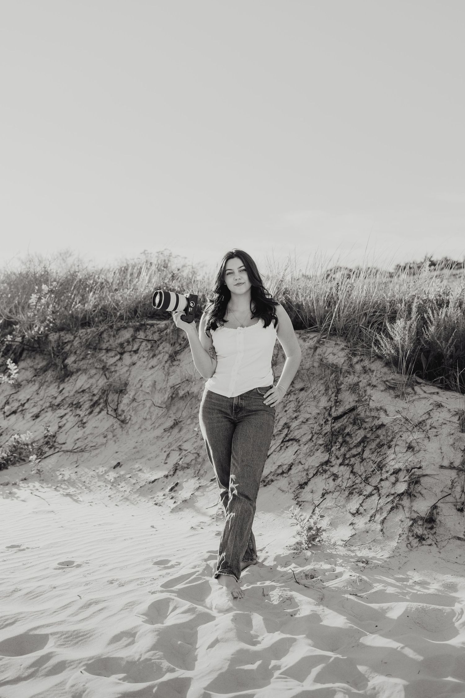 Black and white photo of photographer posing with her camera by the sand dunes editorial style at Horseneck Beach in Westport, Massachusetts