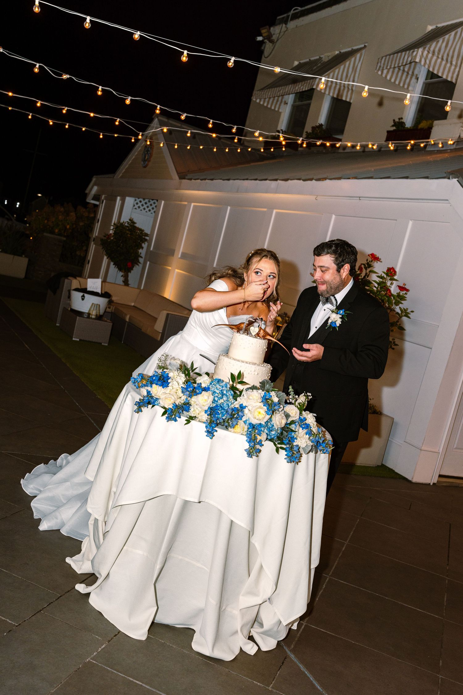 A couple shares a playful moment while eating wedding cake together under twinkling string lights at an outdoor reception in Newport RI