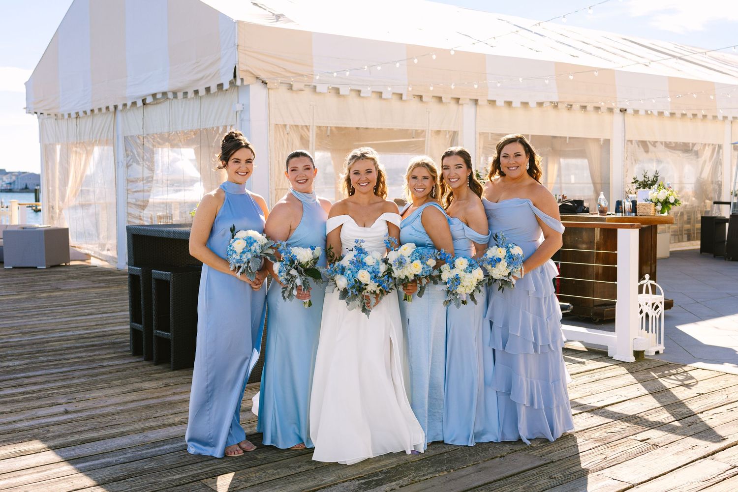 Wedding party wearing light blue bridesmaid dresses poses together on white steps during sunny outdoor celebration at The Regatta Place RI