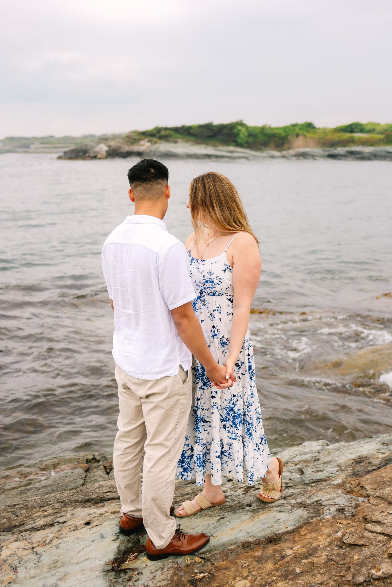 Couple holding hands facing the ocean on rocky shore for engagement photos on Ocean Drive in Newport Rhode Island after surprise proposal