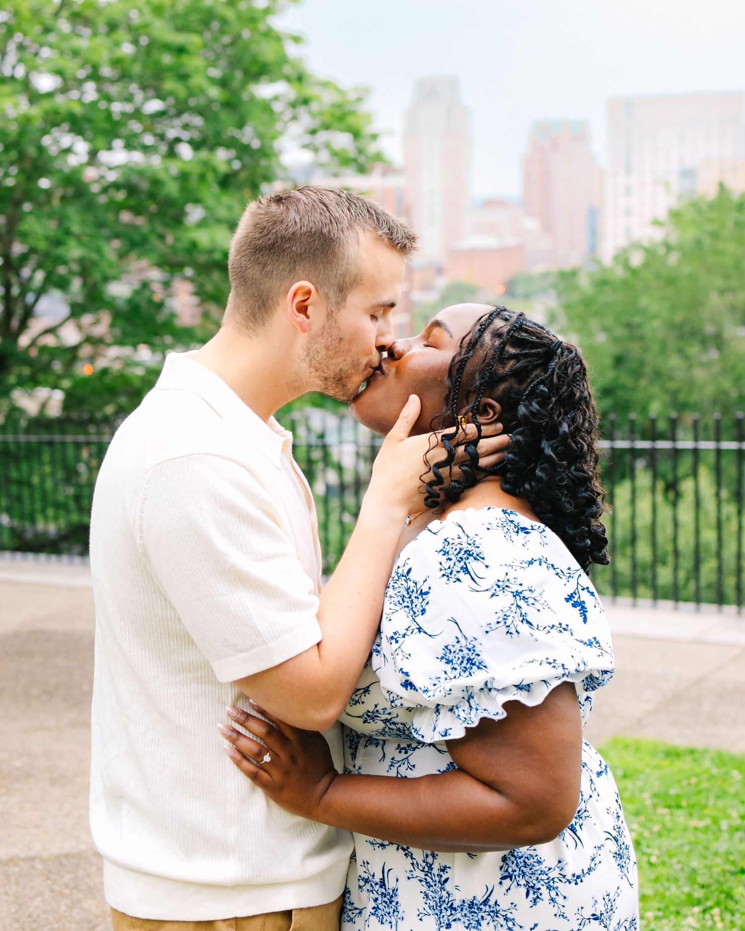 A couple embraces tenderly in an outdoor park setting with green foliage in the background by the Providence city skyline in Rhode Island