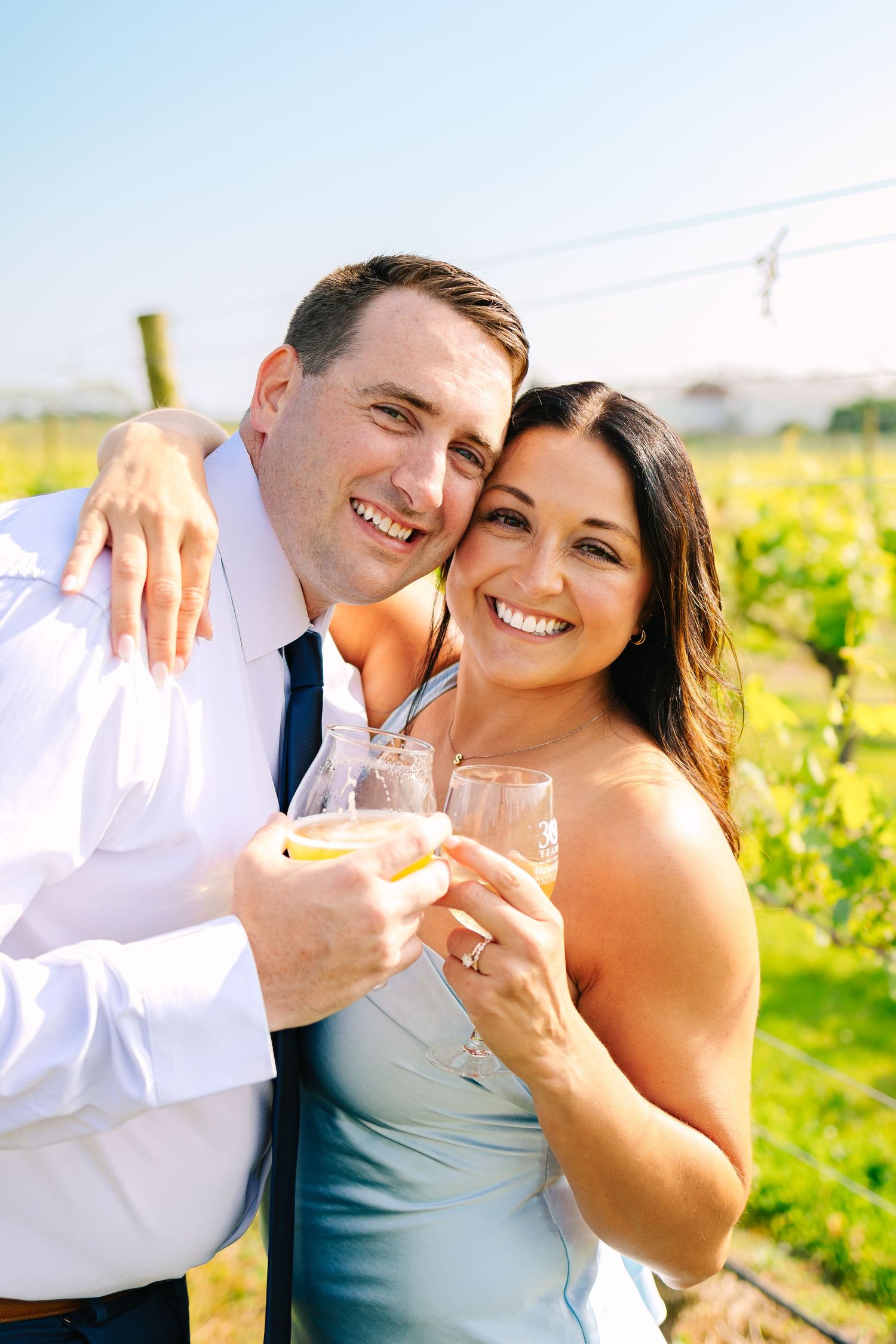 A couple enjoying wine together at a sunny vineyard, dressed in formal attire and smiling warmly.