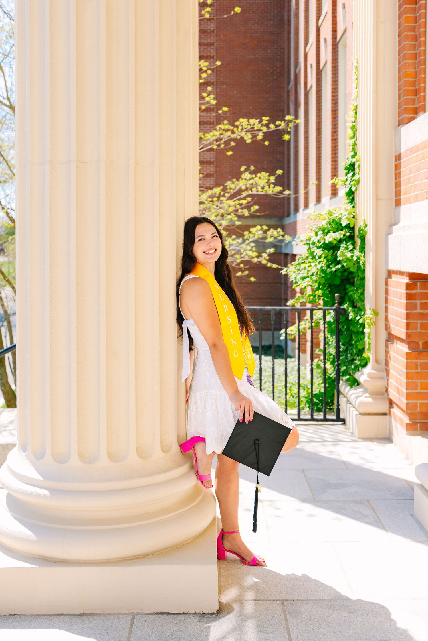 A college grad poses against a white column for graduation photos holding her cap at Worcester State University in Worcester Massachusetts