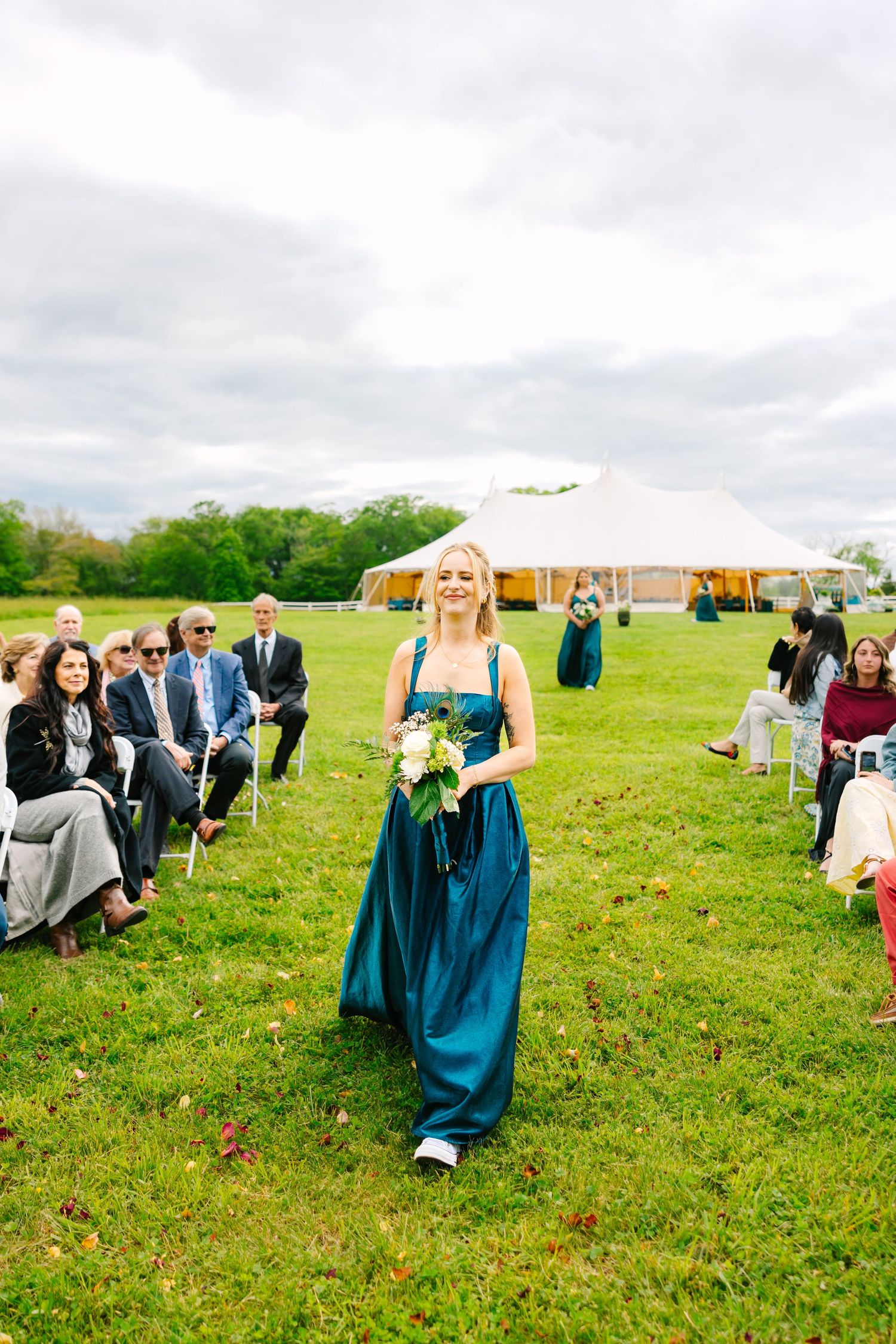 Outdoor wedding ceremony setup in a green field with guests seated in rows and teal bridesmaid dresses visible.