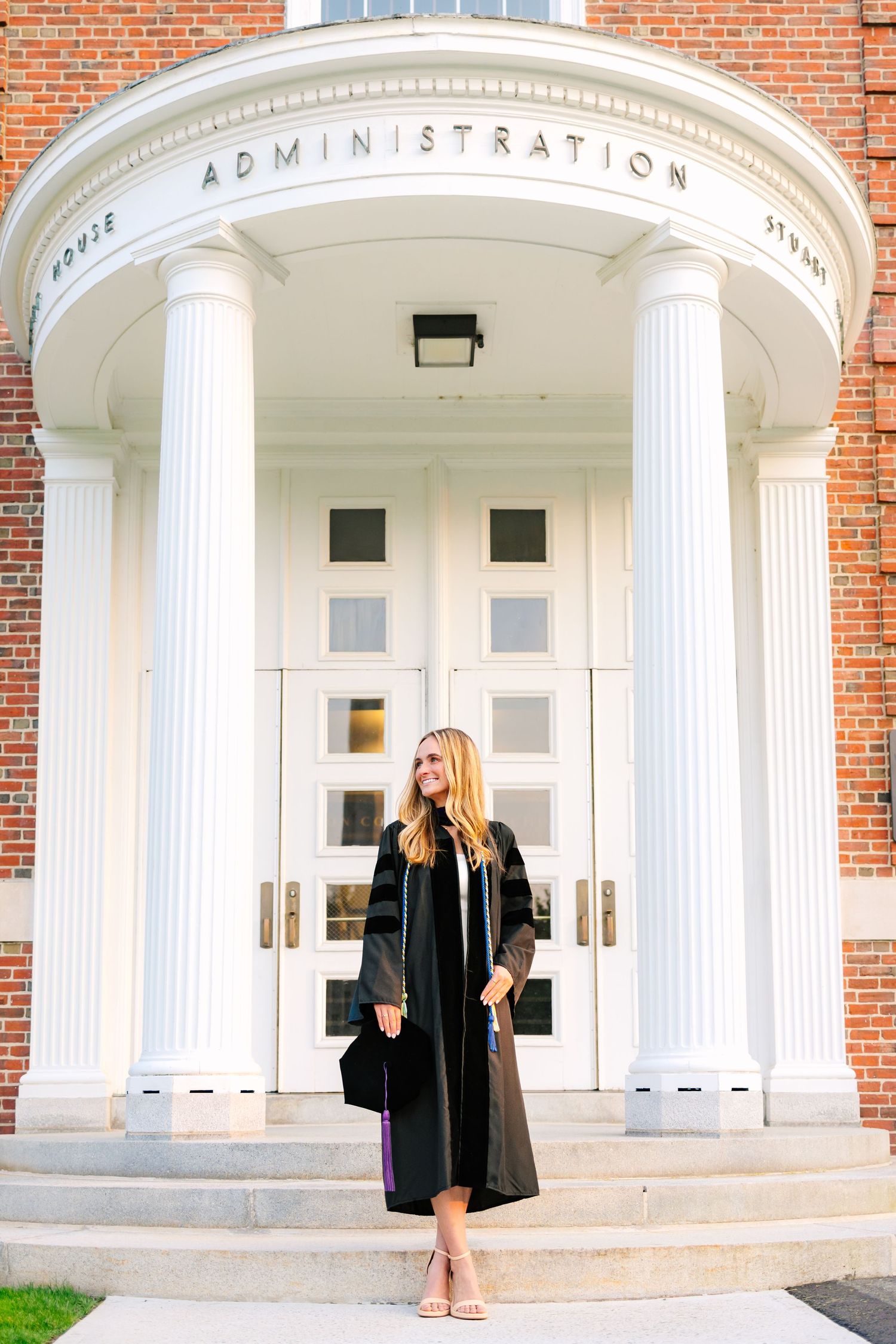 College grad stands in front of a brick building with white columns wearing a cap and gown for graduation photos at Boston College Law