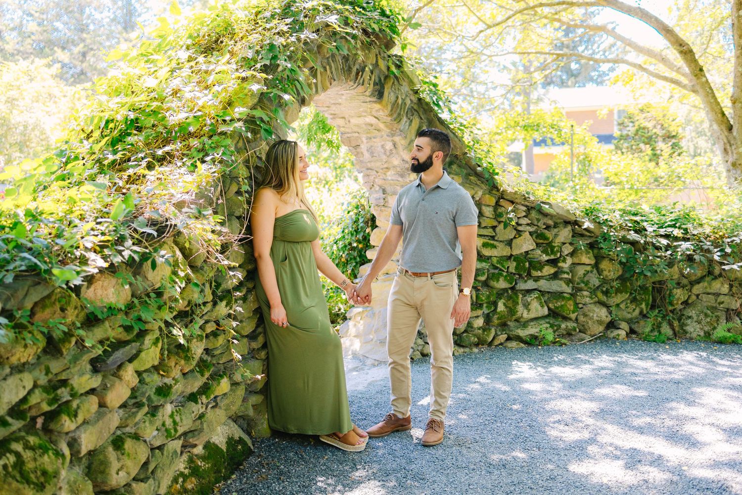 Two people walk through a sunlit garden path during engagement photos in summertime at Blithewold Mansion in Bristol Rhode Island