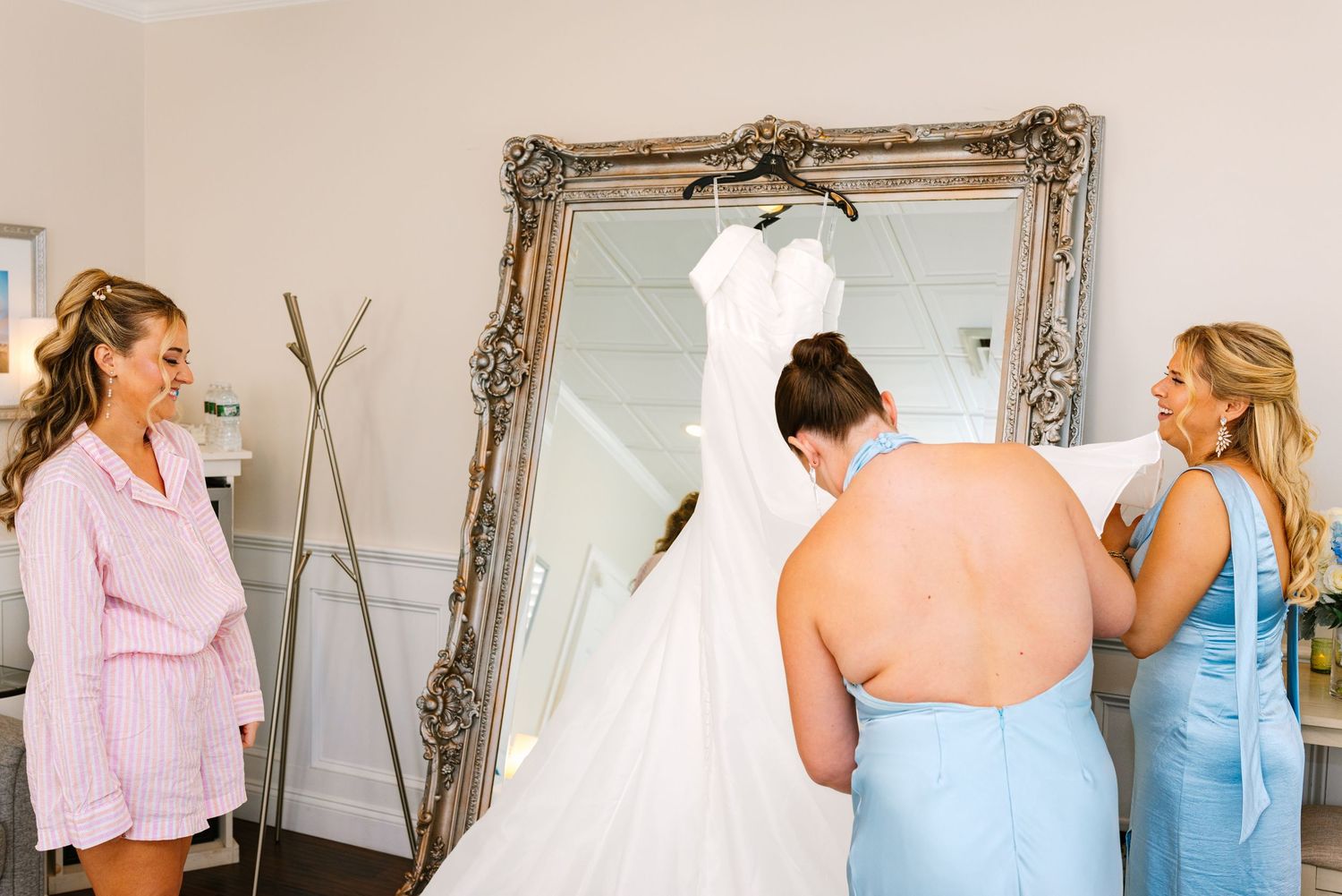 People in a bridal boutique admire a white wedding dress displayed in an ornate silver frame mirror.