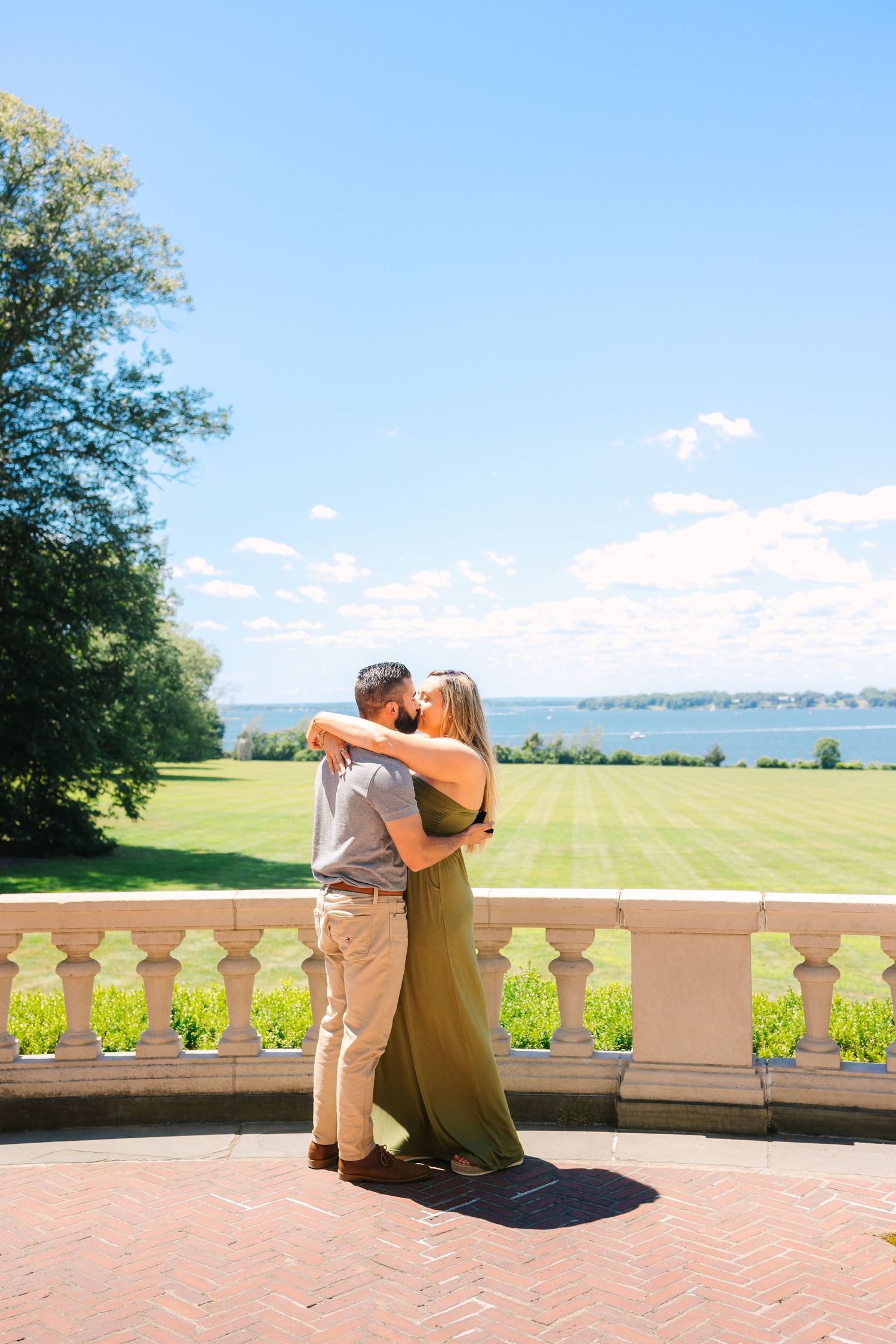 A couple shares a romantic moment on a brick terrace overlooking scenic waterfront grounds on a sunny summer day.