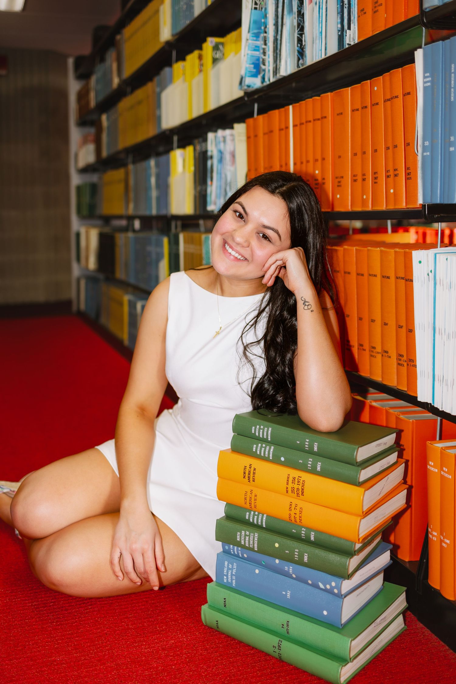 A college grad posing with stacked books on red carpet in a library aisle for graduation photos at UMASS Dartmouth Massachusetts