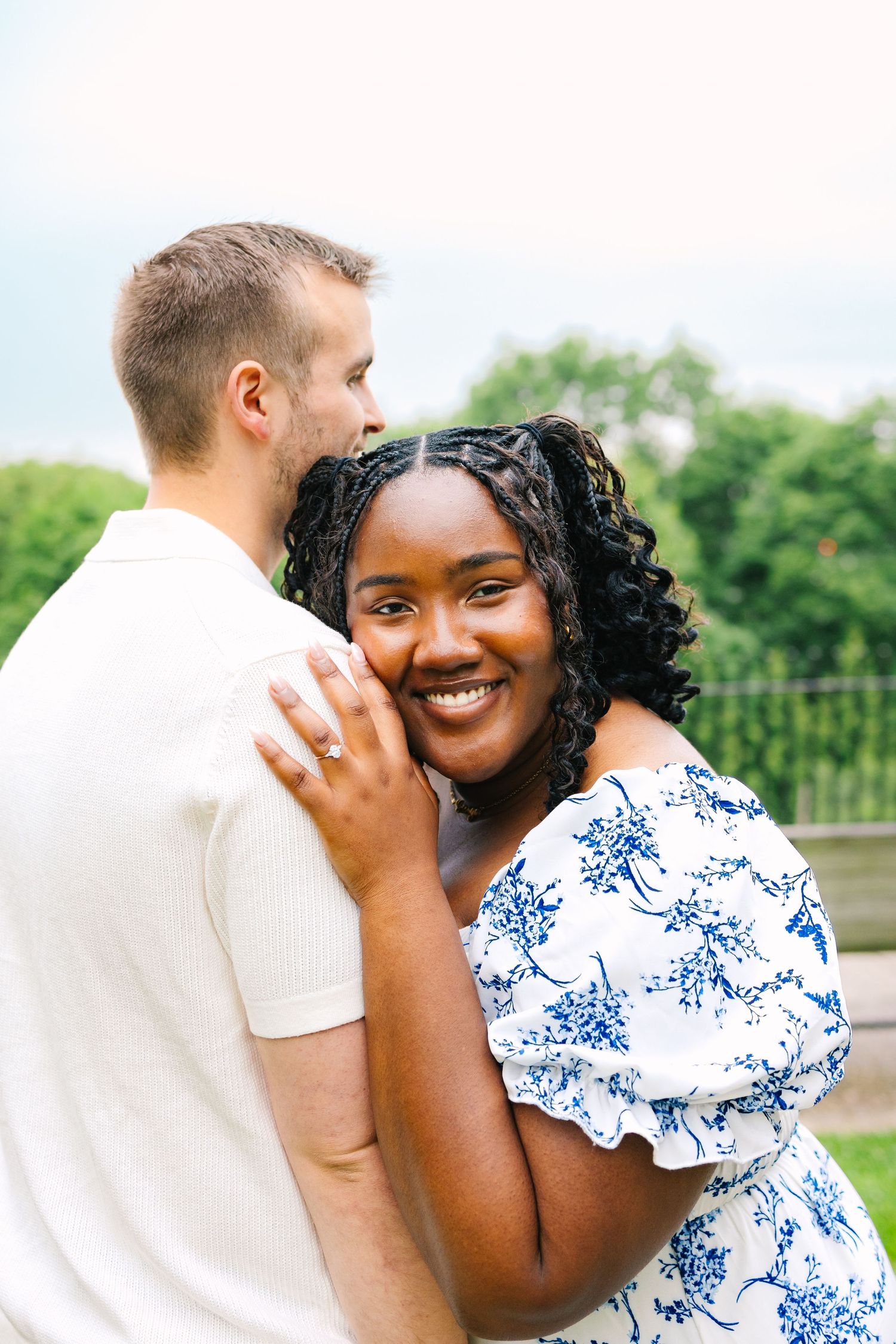 A couple embraces outdoors for summertime engagement photos on a cloudy day at a park with a view of the Providence city skyline in RI