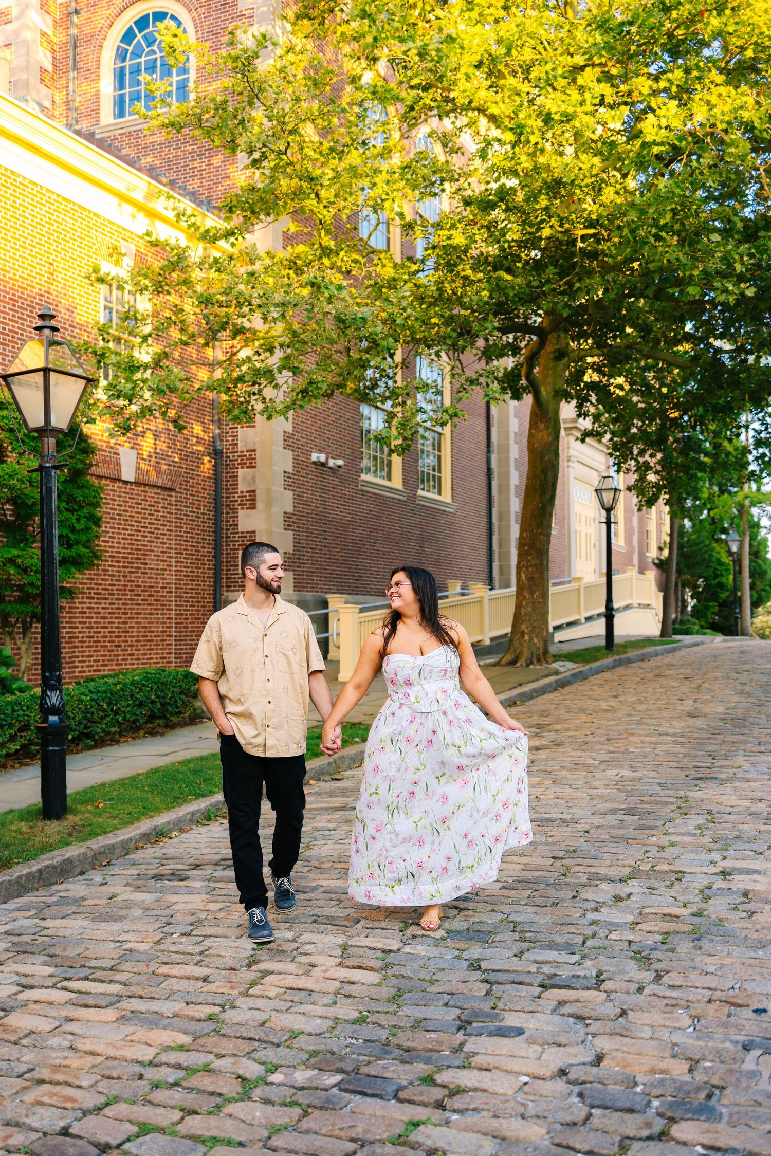 A couple strolls along a picturesque cobblestone street with brick buildings and autumn trees in historic New Bedford, MA.
