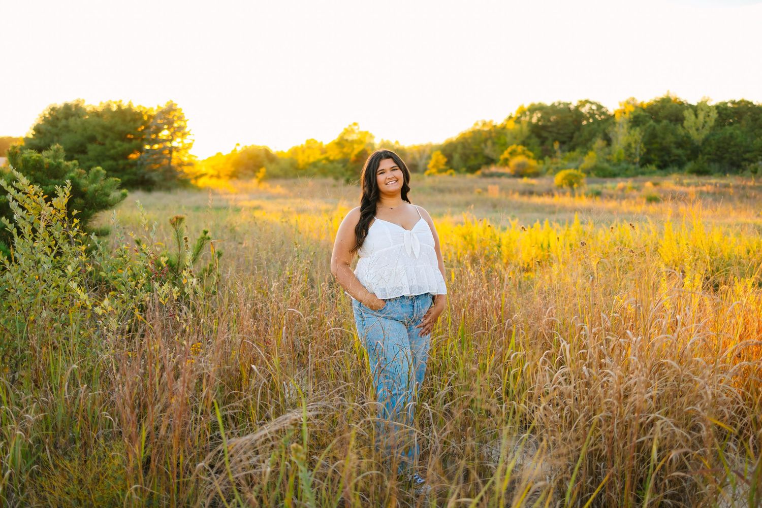 A high school senior stands in a golden field of tall grass at sunset creating a dreamy autumn scene for senior photos in West Greenwich RI