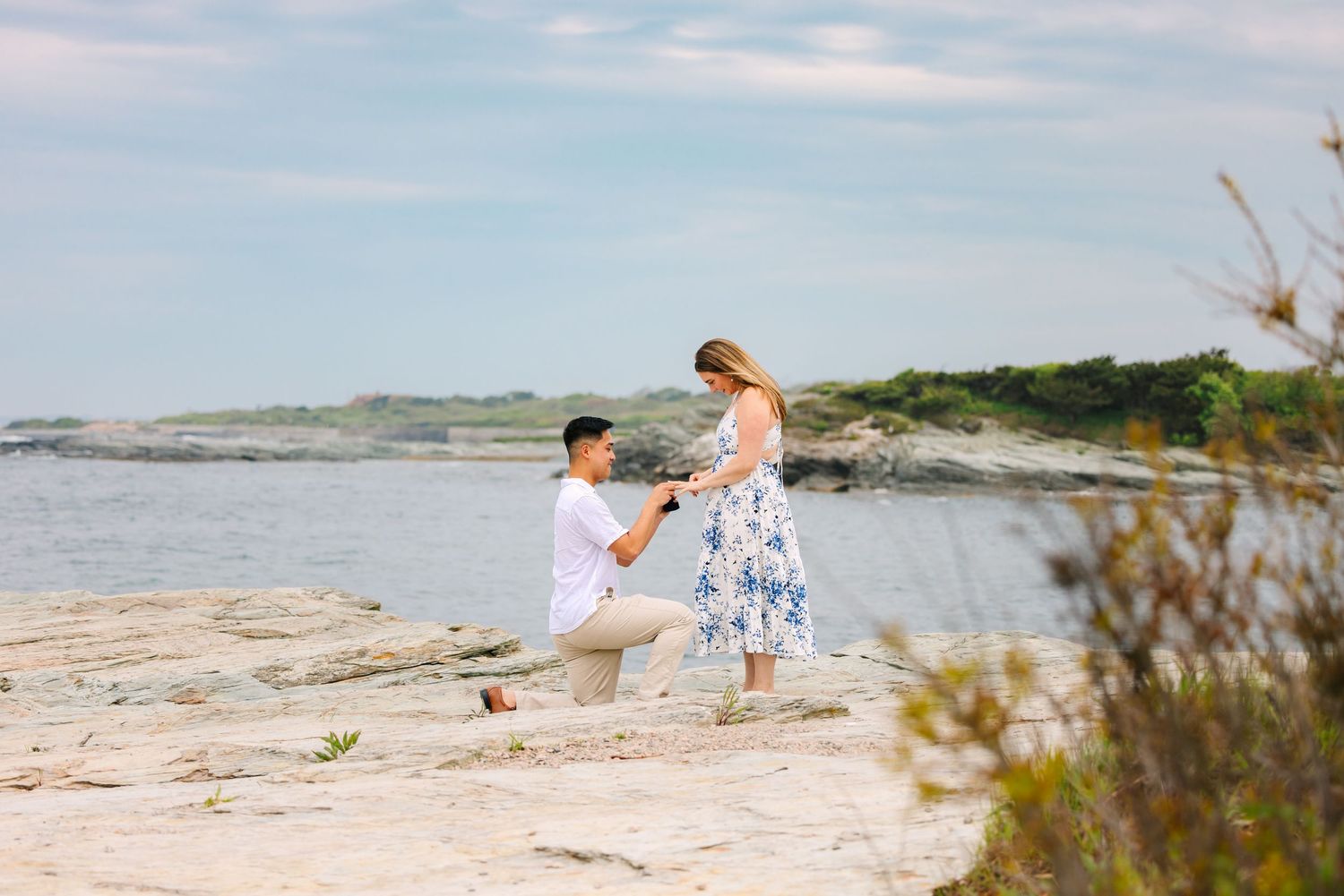 Man proposes to woman on rocky beach shoreline on Ocean Drive in Newport Rhode Island, both dressed in summer attire with ocean backdrop.