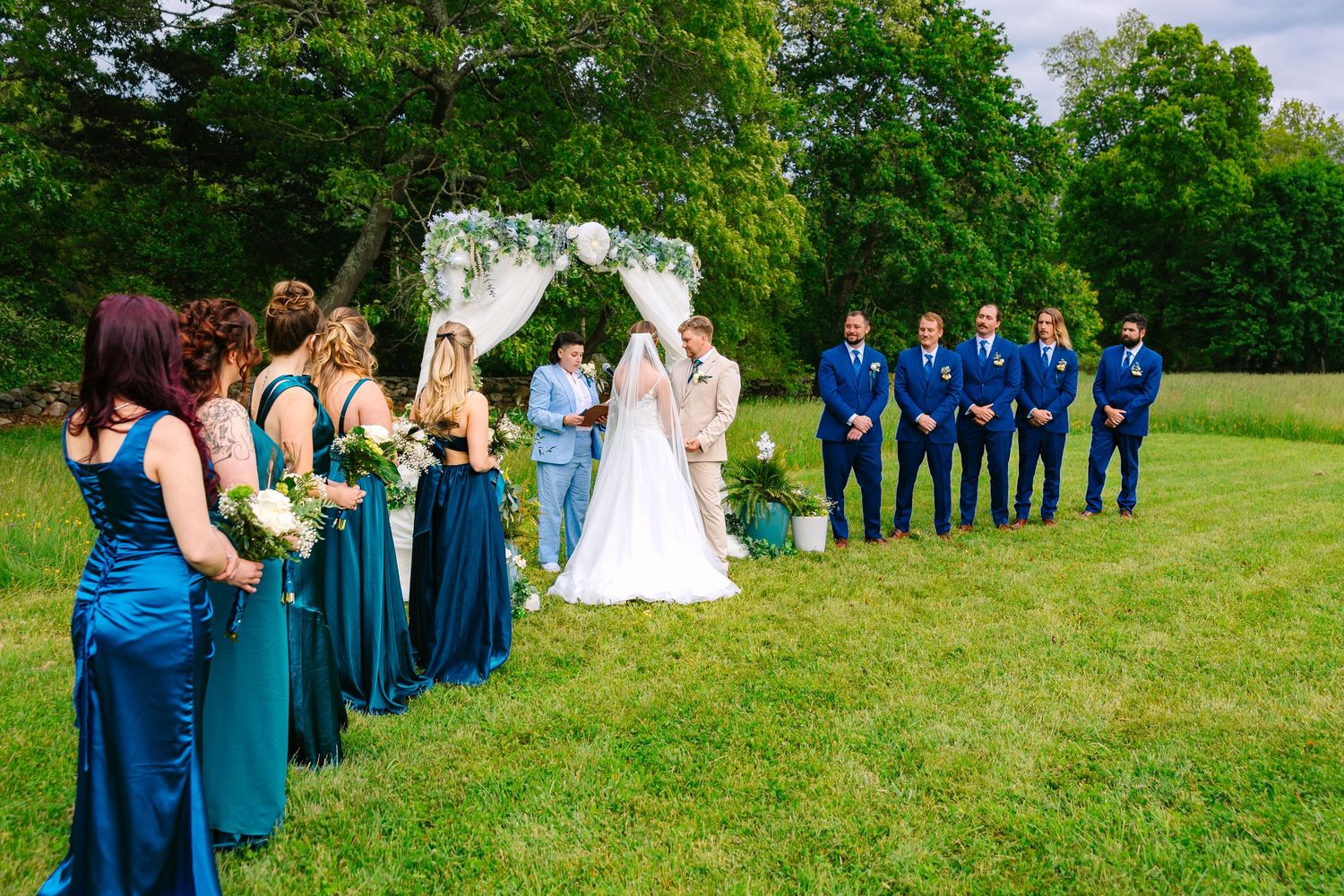 Outdoor wedding ceremony setup with wedding party in blue and teal attire standing on opposite sides of floral arch.