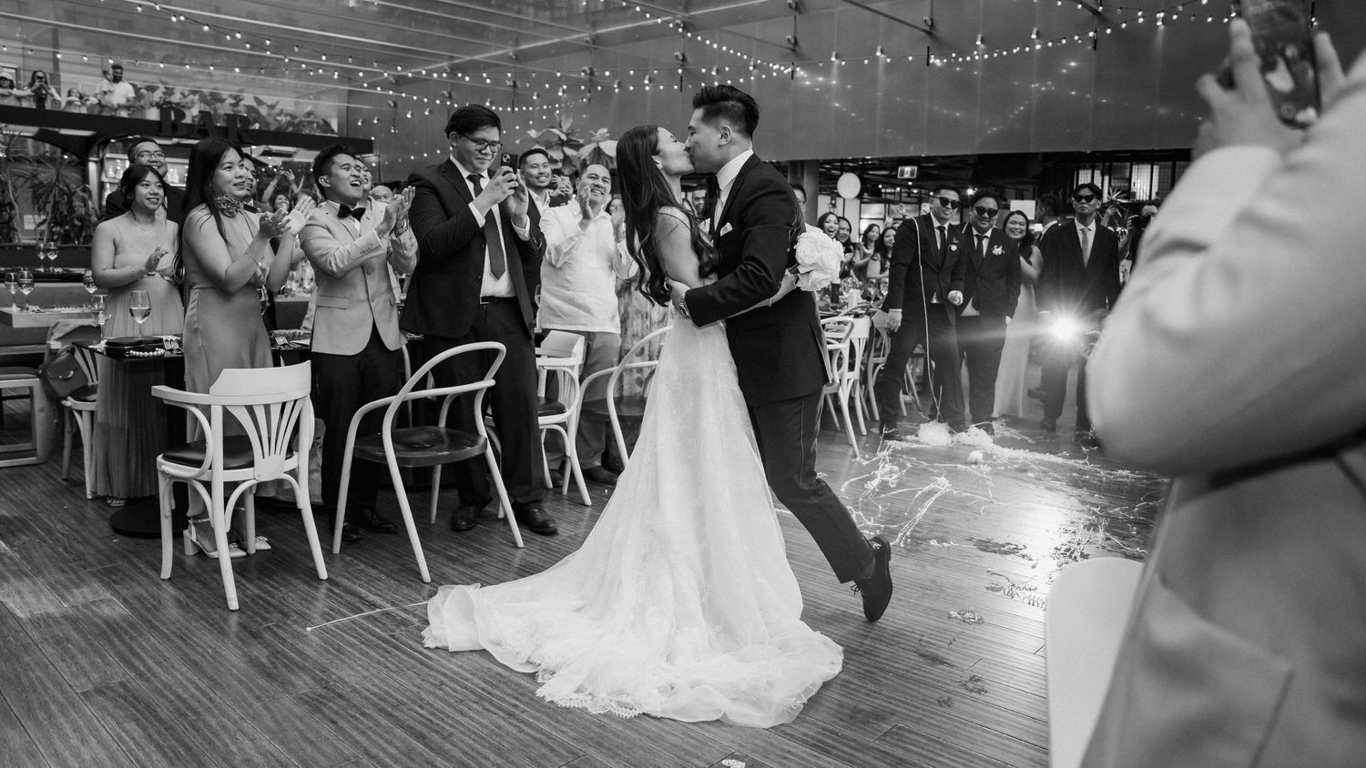 Couple shares intimate first dance under string lights in rustic venue while guests look on.