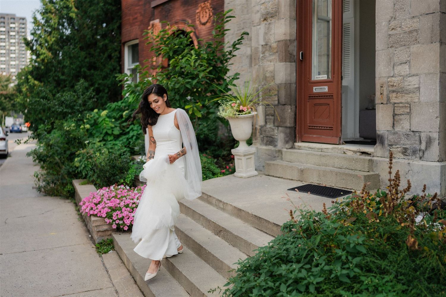 Elegant bride in flowing white dress standing on stone steps surrounded by pink flowers and greenery.