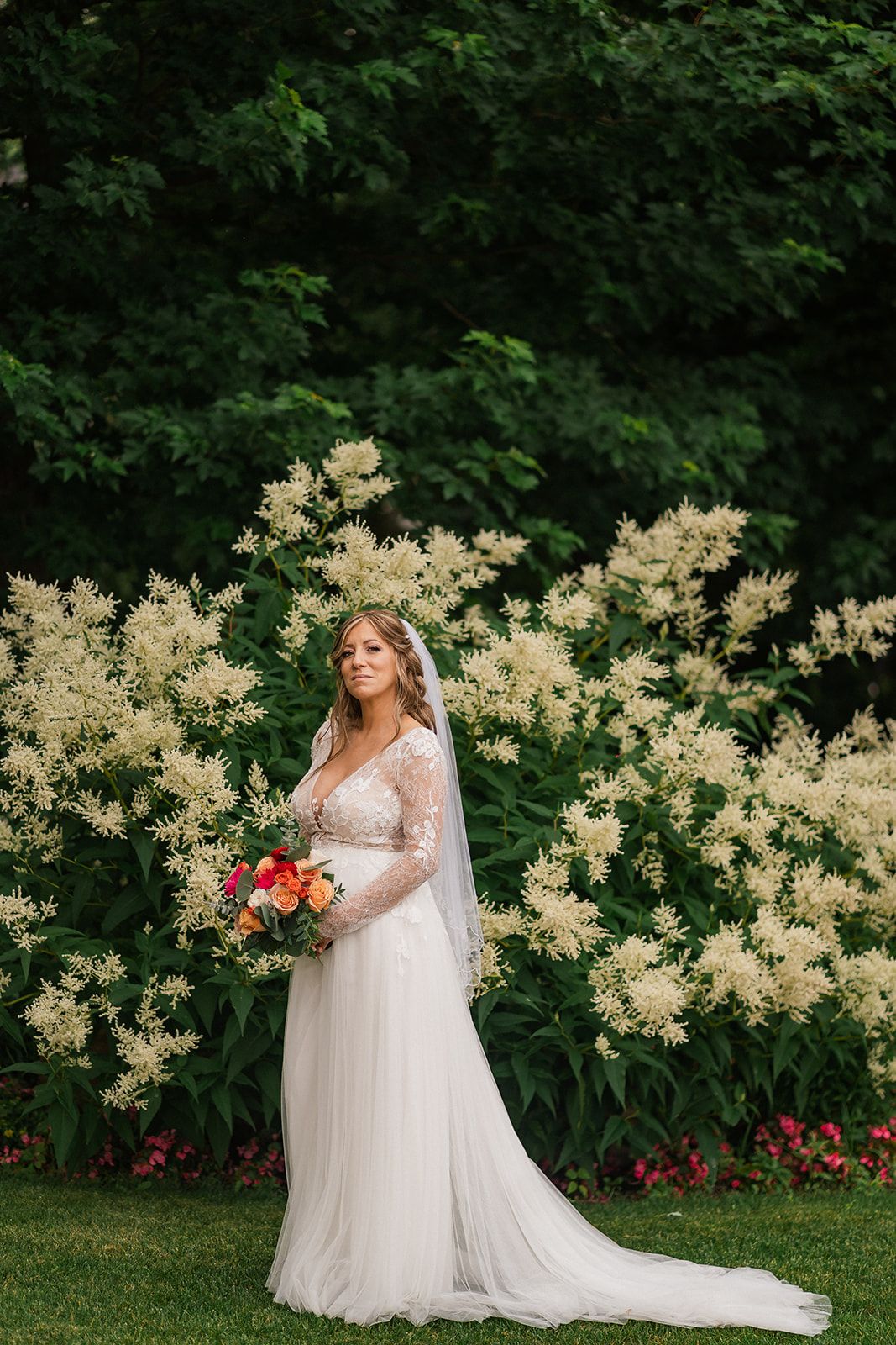 A bride in a flowing gown poses in a garden with blooming white flowers and red tulips.