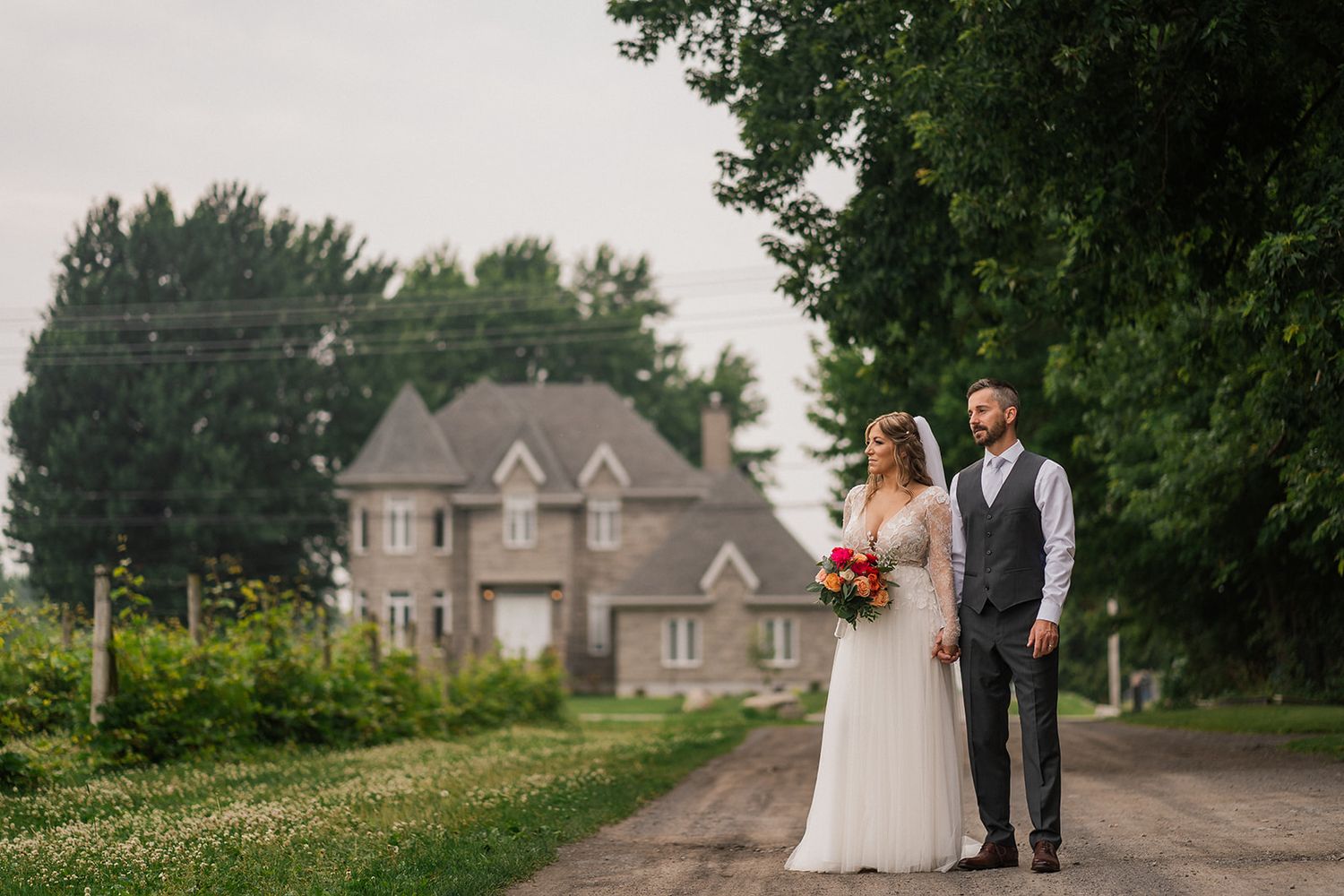 A wedding couple poses on a tree-lined path with a stone house in the background.
