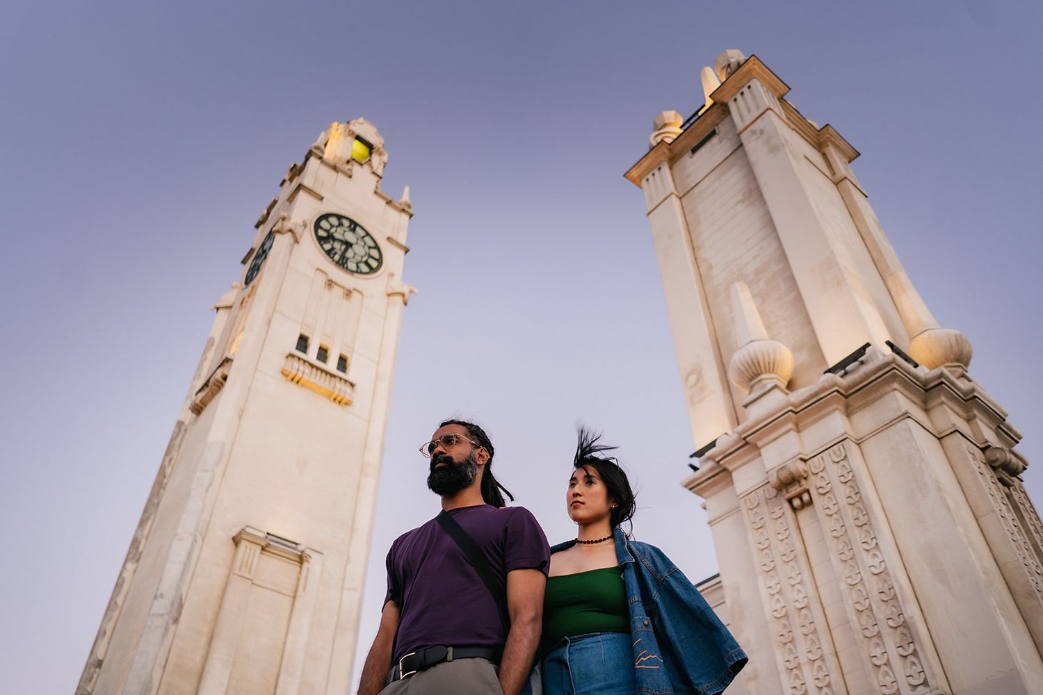Couple poses against tall clock towers at sunset wearing casual attire.