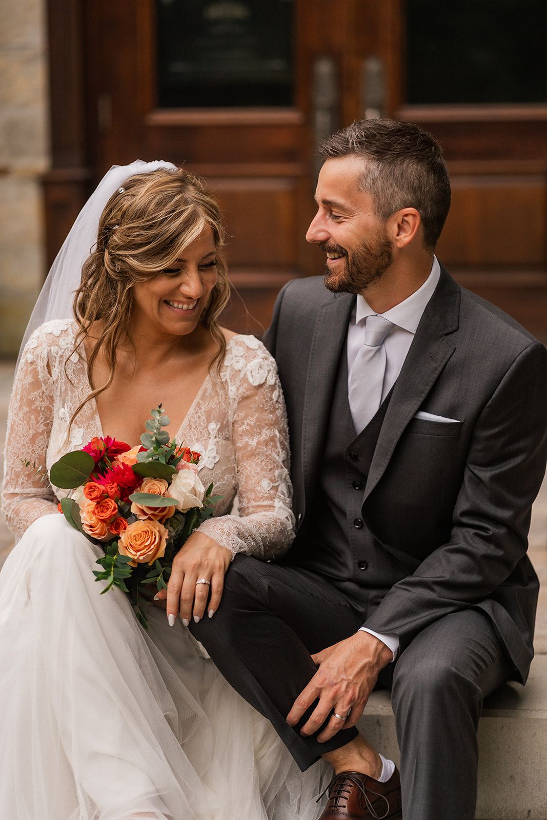 A happy bride and groom share laughter while sitting together on their wedding day.