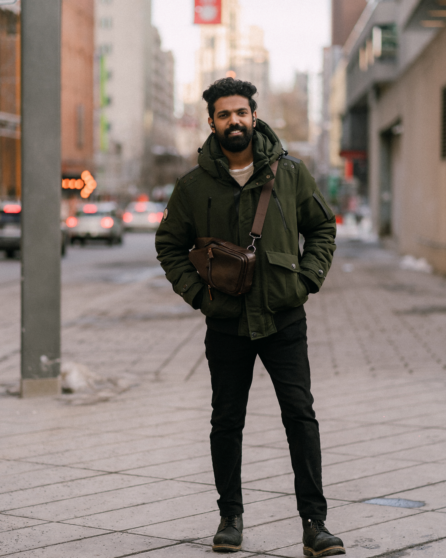 Person in a dark winter coat and messenger bag standing on an urban sidewalk in a city setting.
