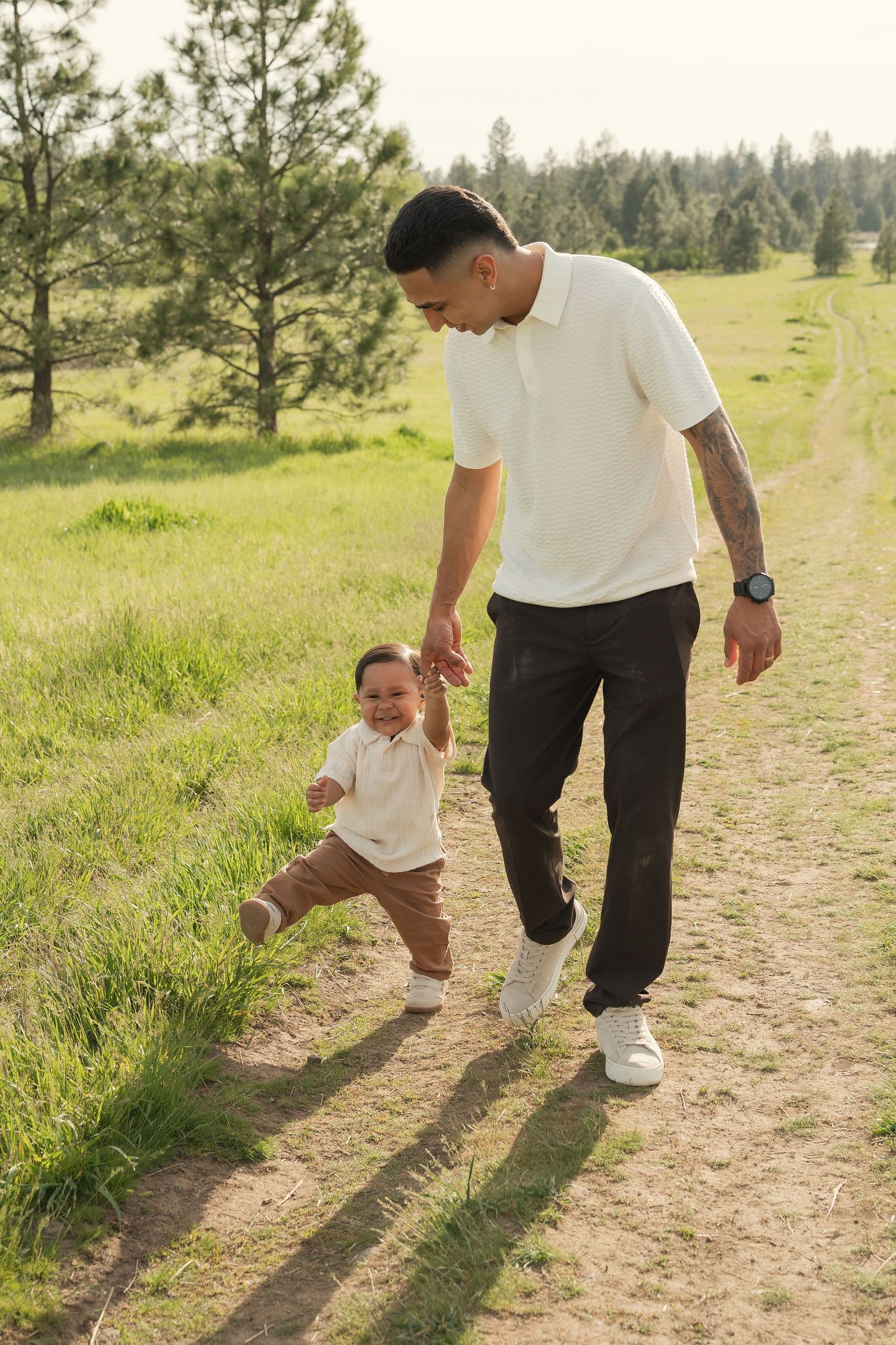 Father and toddler son walking hand in hand on a sunny meadow path surrounded by green trees.