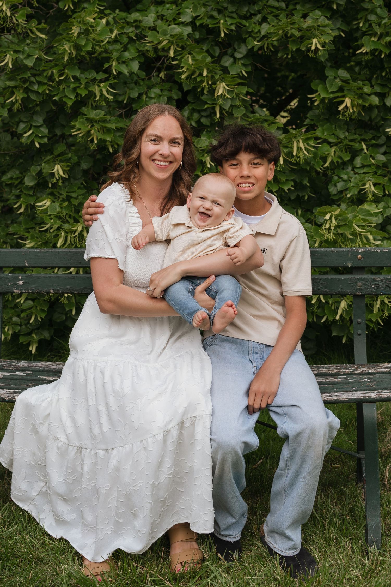 A smiling woman in white dress sits on a bench with a laughing baby and teenage boy outdoors.