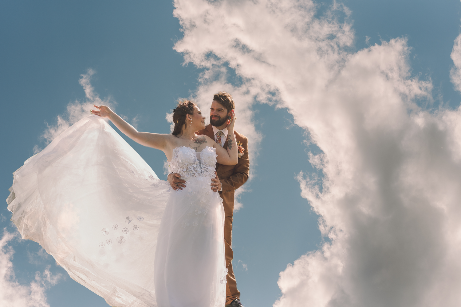 Bride in flowing white dress and groom embrace against blue sky with clouds in romantic wedding photo.