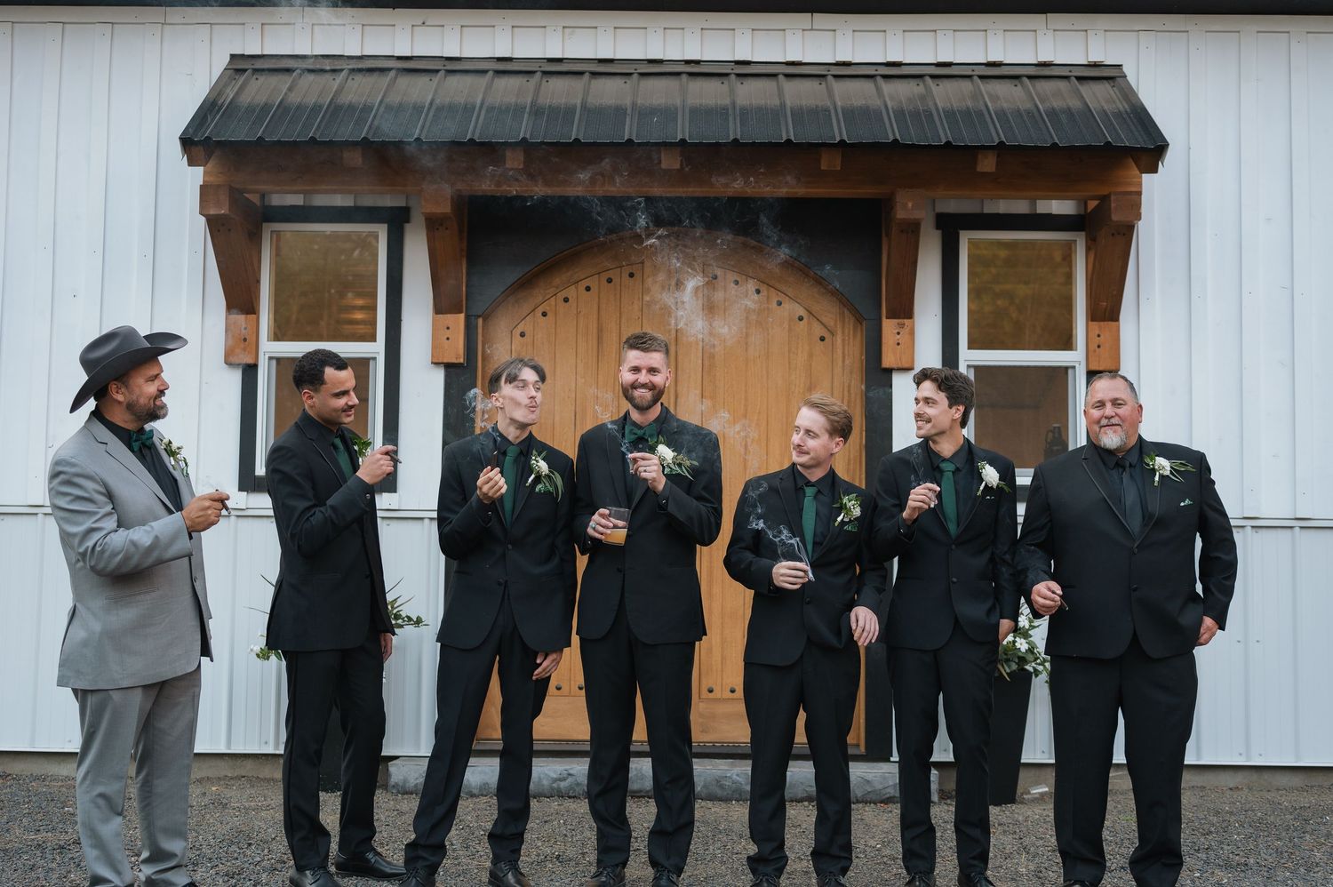 Groomsmen in black suits with green boutonnieres laughing and smoking cigars outside a white barn wedding venue.