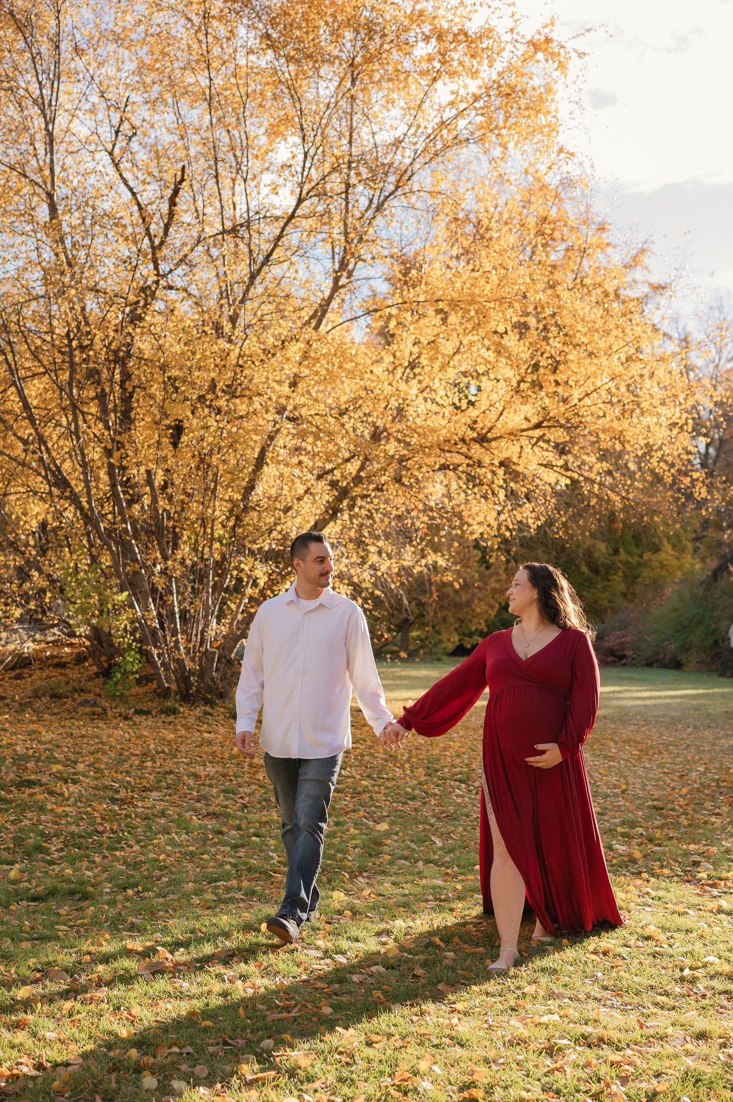 Pregnant couple holding hands walking in autumn park, woman in red maternity gown, golden fall foliage background.