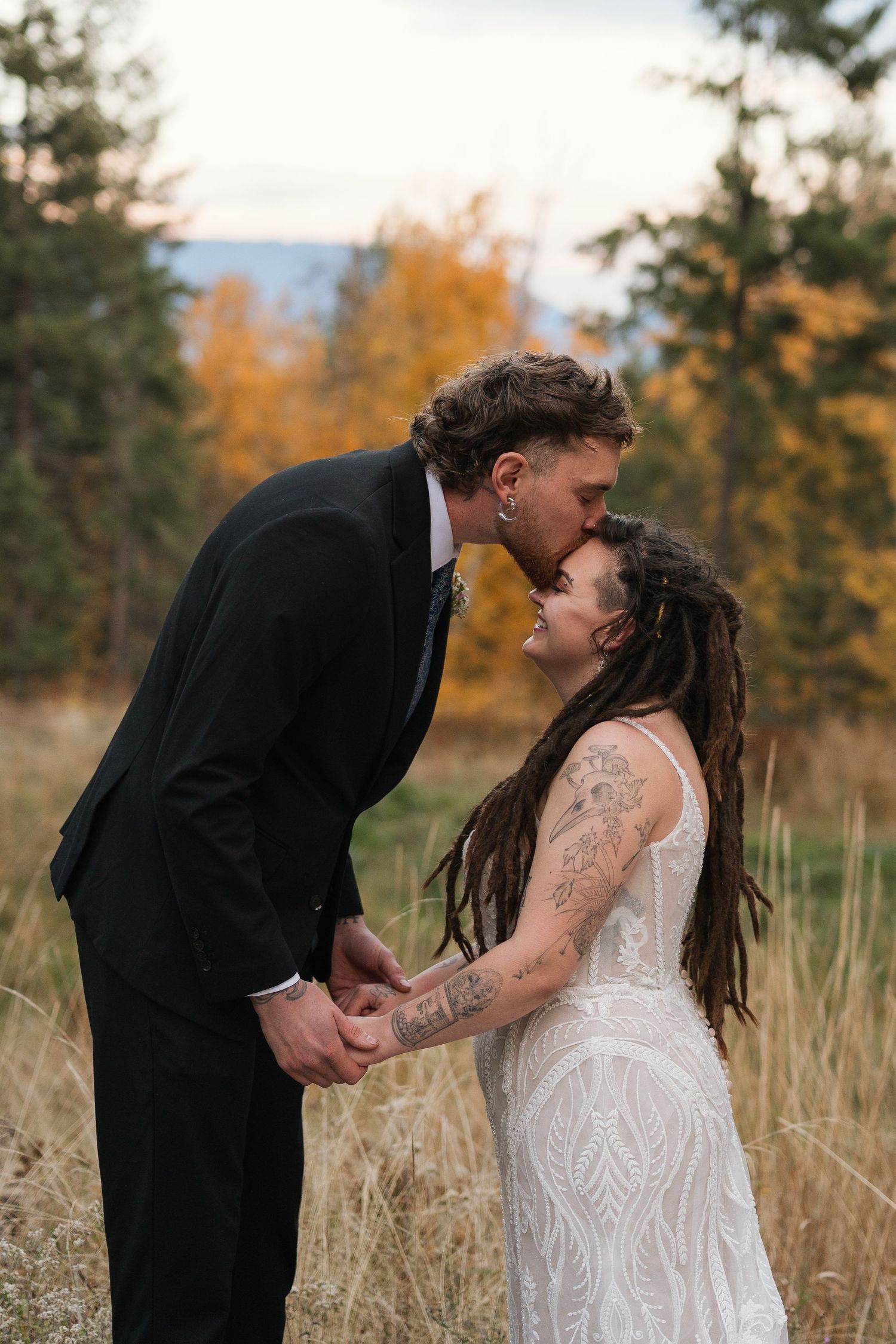 A romantic wedding couple shares intimate kisses in a golden autumn field with evergreen trees in the background.