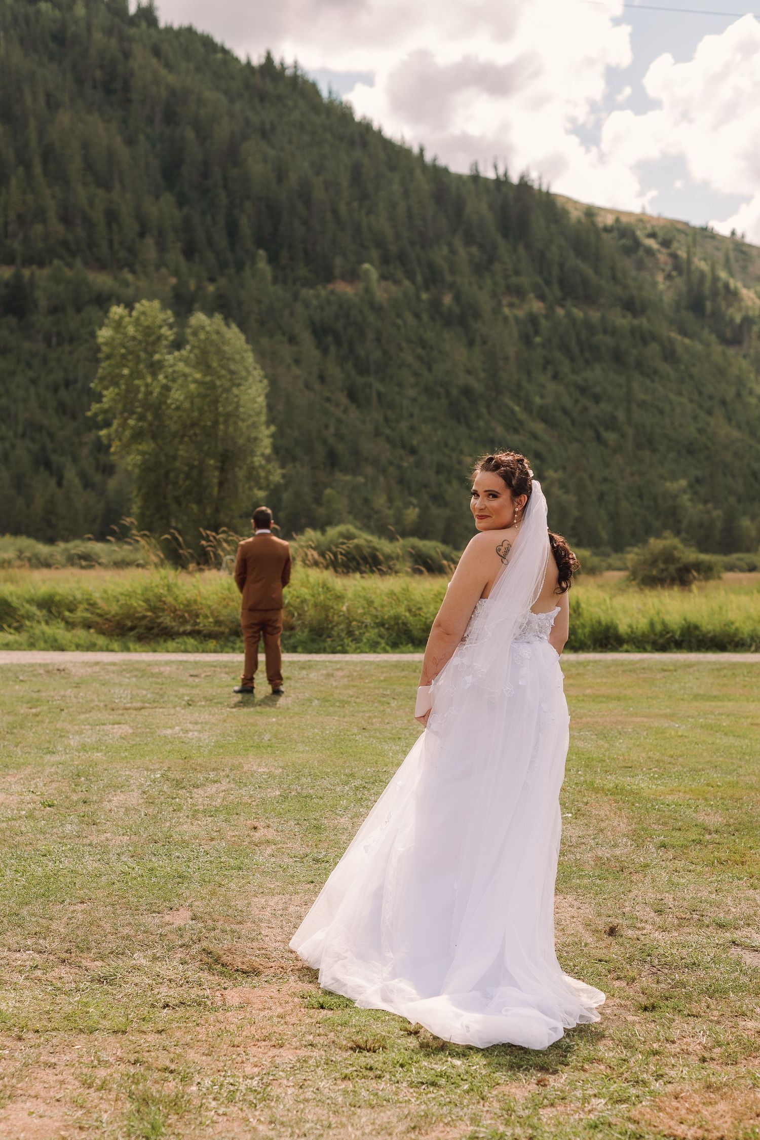 Bride in white gown looks back at camera while groom stands in distance on green meadow with mountain backdrop.
