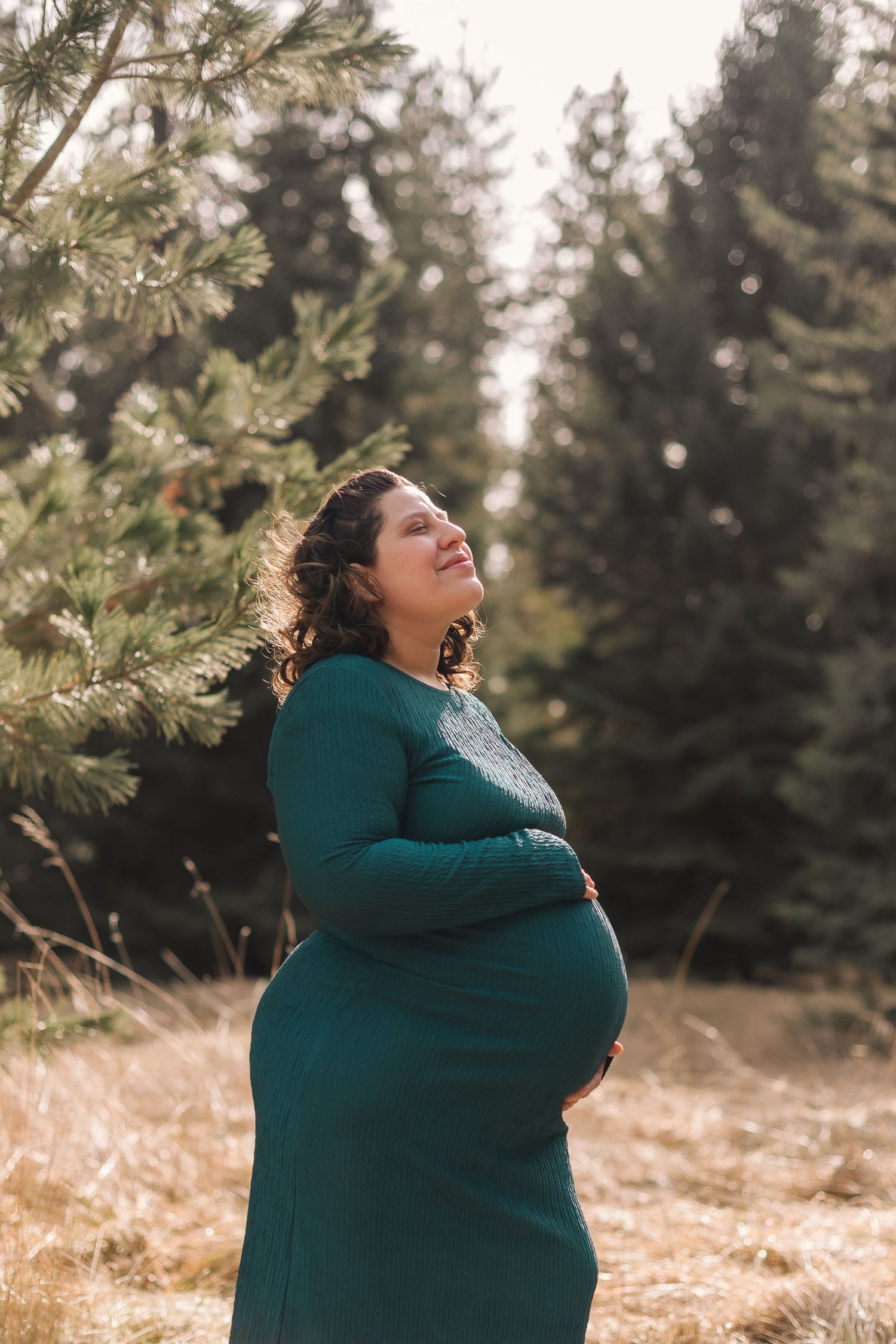 A person in a teal sweater stands in a field of dried grass with evergreen trees in the background on a sunny day.