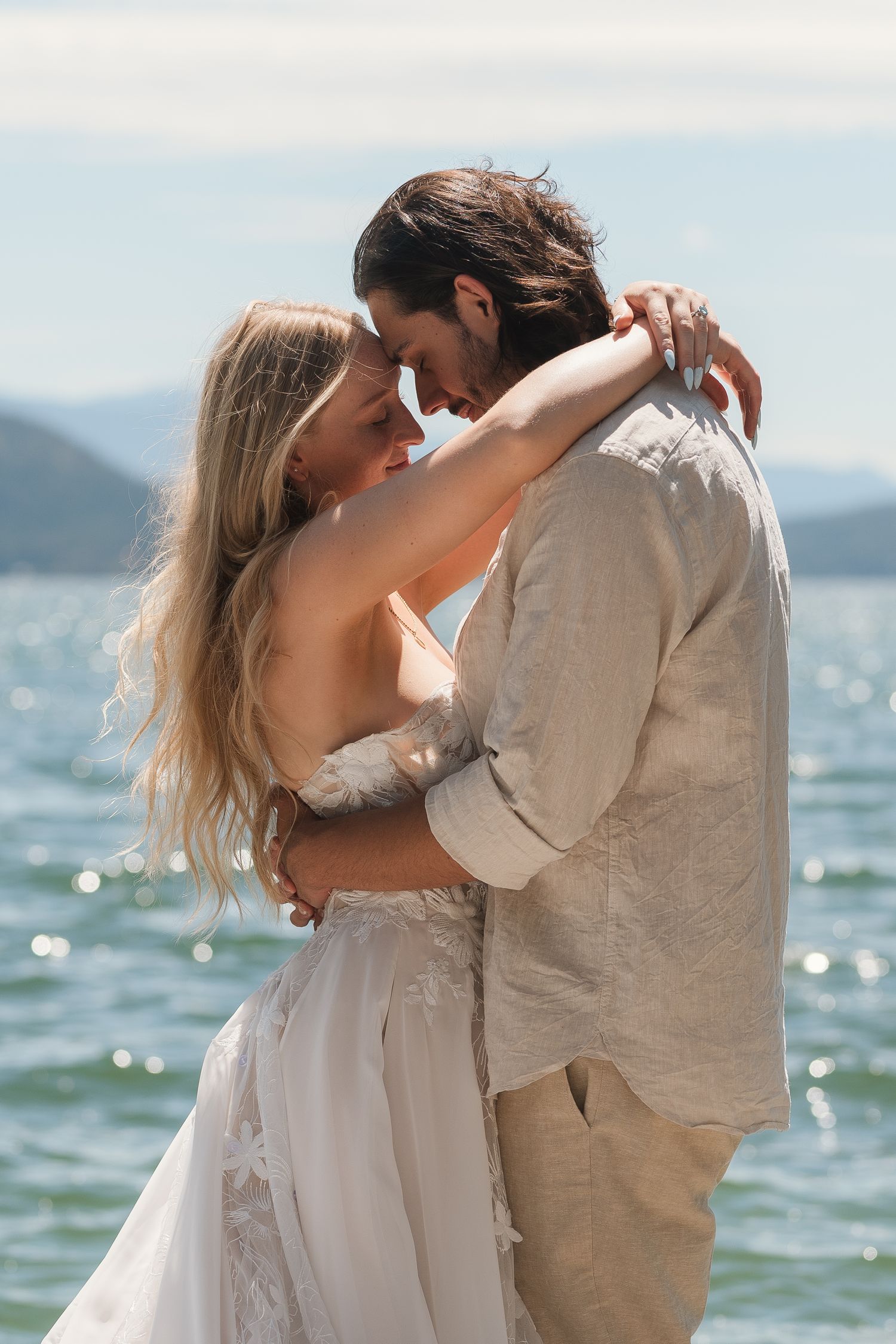 A couple in bohemian wedding attire embracing romantically by a sparkling ocean with mountain views.