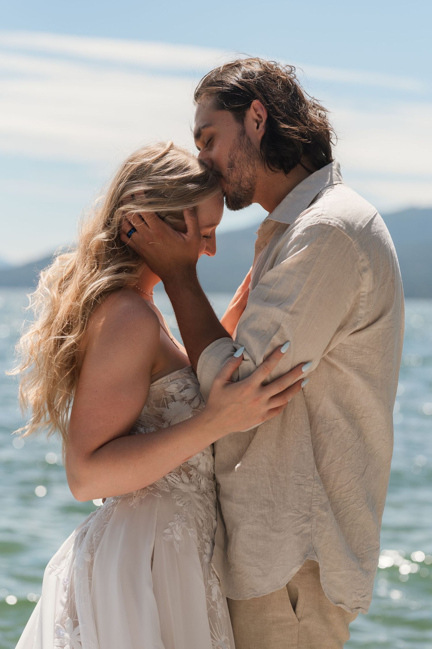 Couple sharing a romantic moment by the ocean, bride in white lace dress and groom in linen shirt at beach wedding.